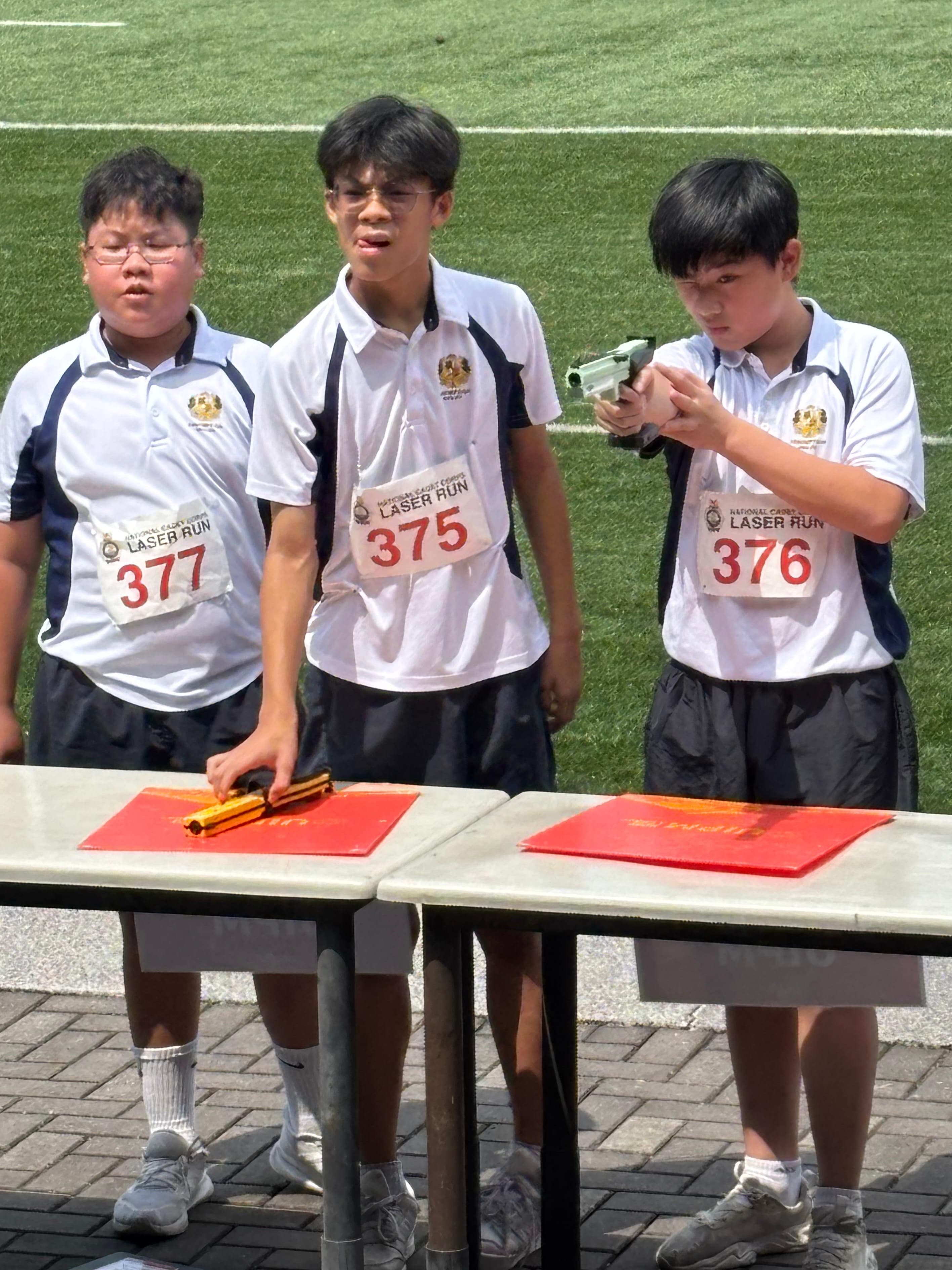 Three boys wearing Laser Run bibs numbered 375, 376, and 377 stand near a table, one aiming a laser pistol.