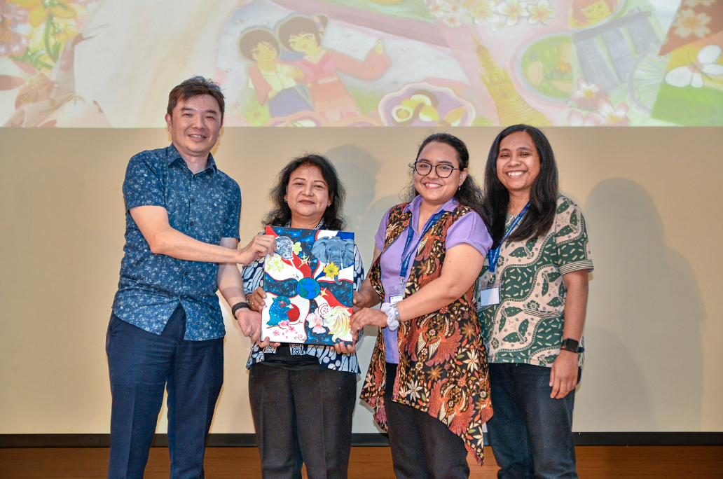 Four people stand together on a stage, smiling, as two of them hold a colorful illustrated book or artwork.