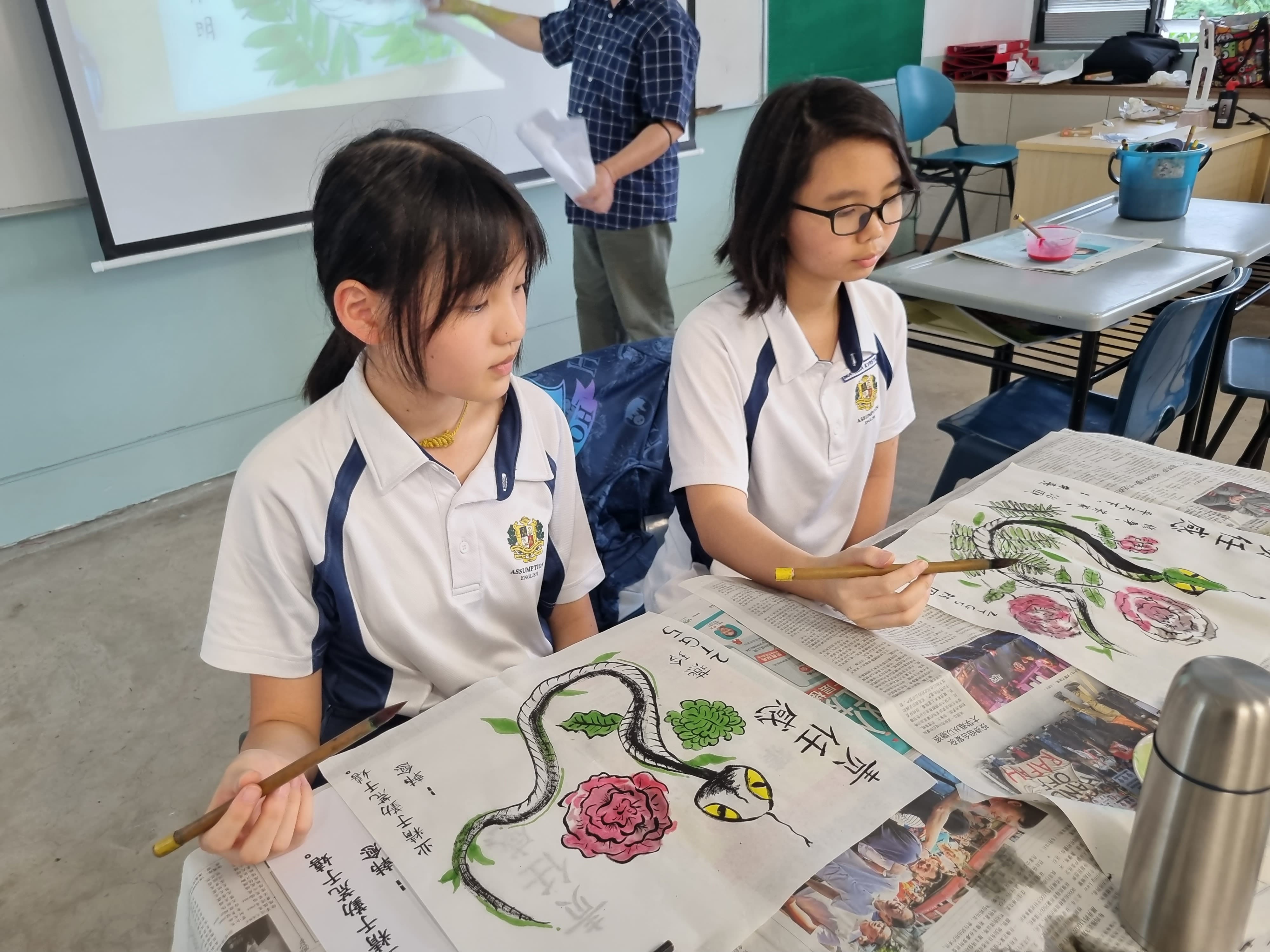 Two students in school uniforms hold paintbrushes over artwork featuring snakes and flowers during an art class.