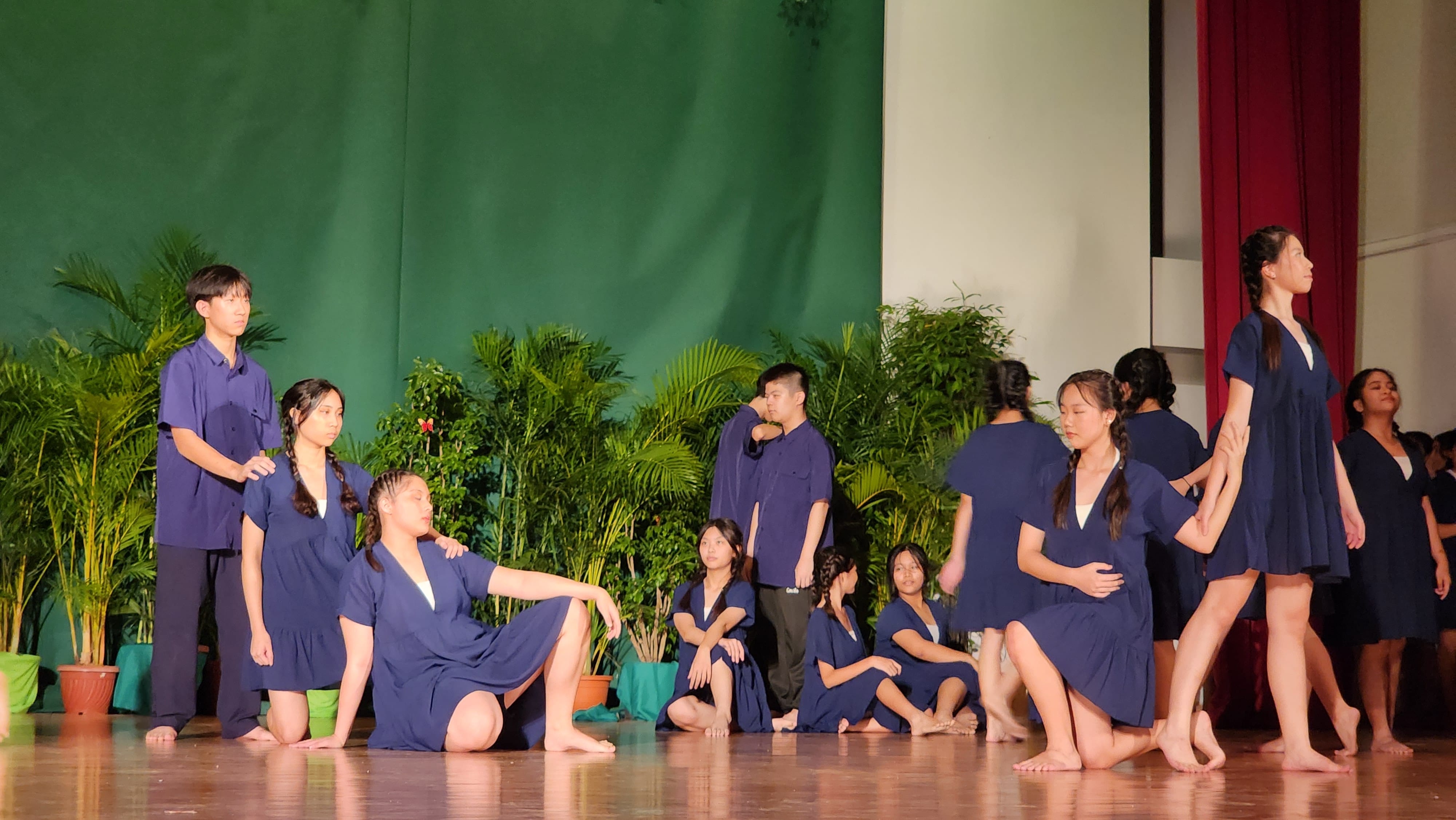 Young performers in navy blue outfits pose on a wooden stage with green plants and a green curtain backdrop.