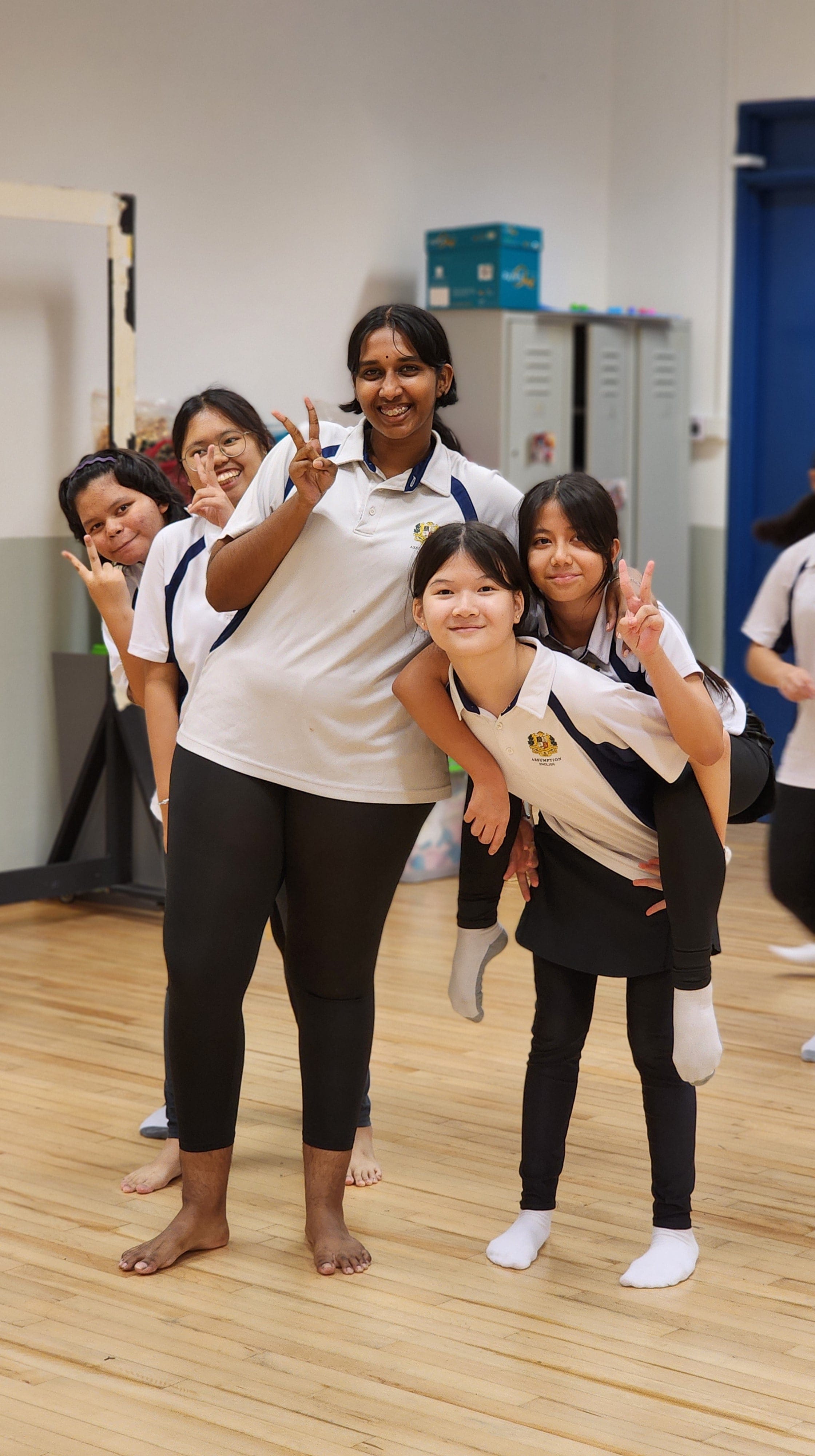 Five teenage girls in white school uniforms pose and smile in a wooden-floored room, making peace signs.