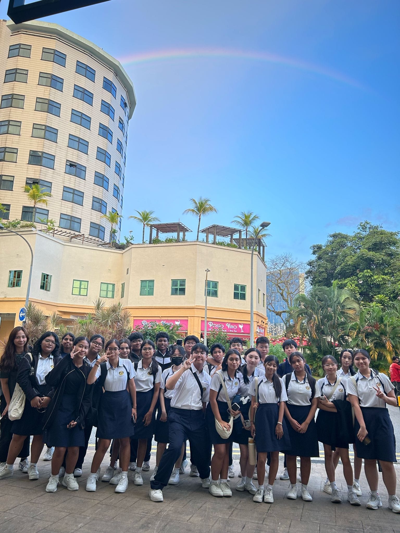 A large group of students in white and navy uniforms pose outdoors near a round building with a rainbow in the sky.
