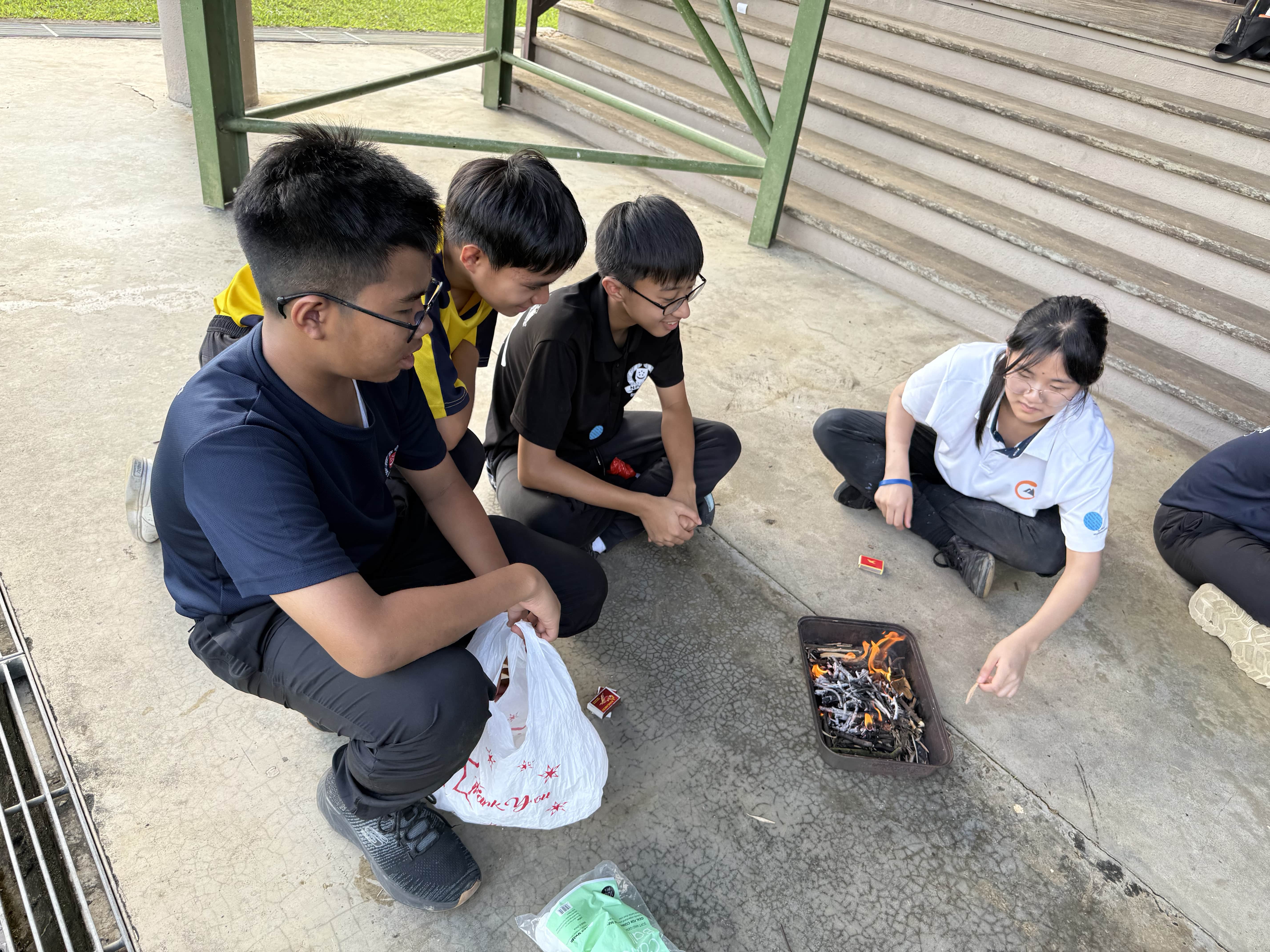Four students sit on concrete around a small tray with burning materials during an outdoor activity.