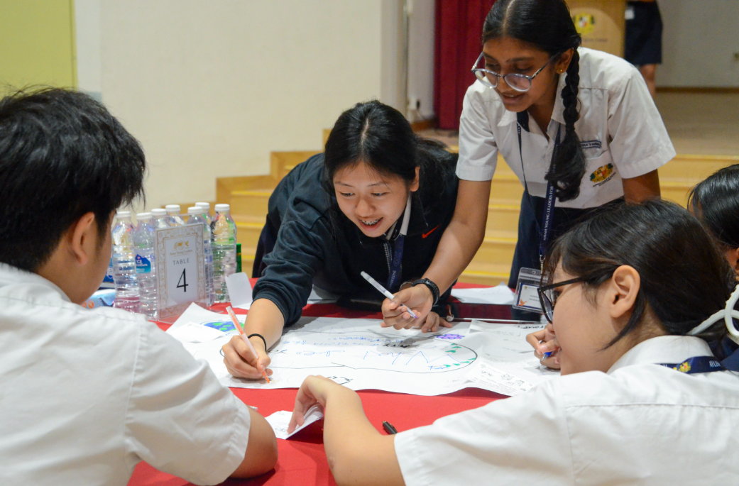 Four school students in uniforms collaborate around a table covered with papers, writing and drawing together.