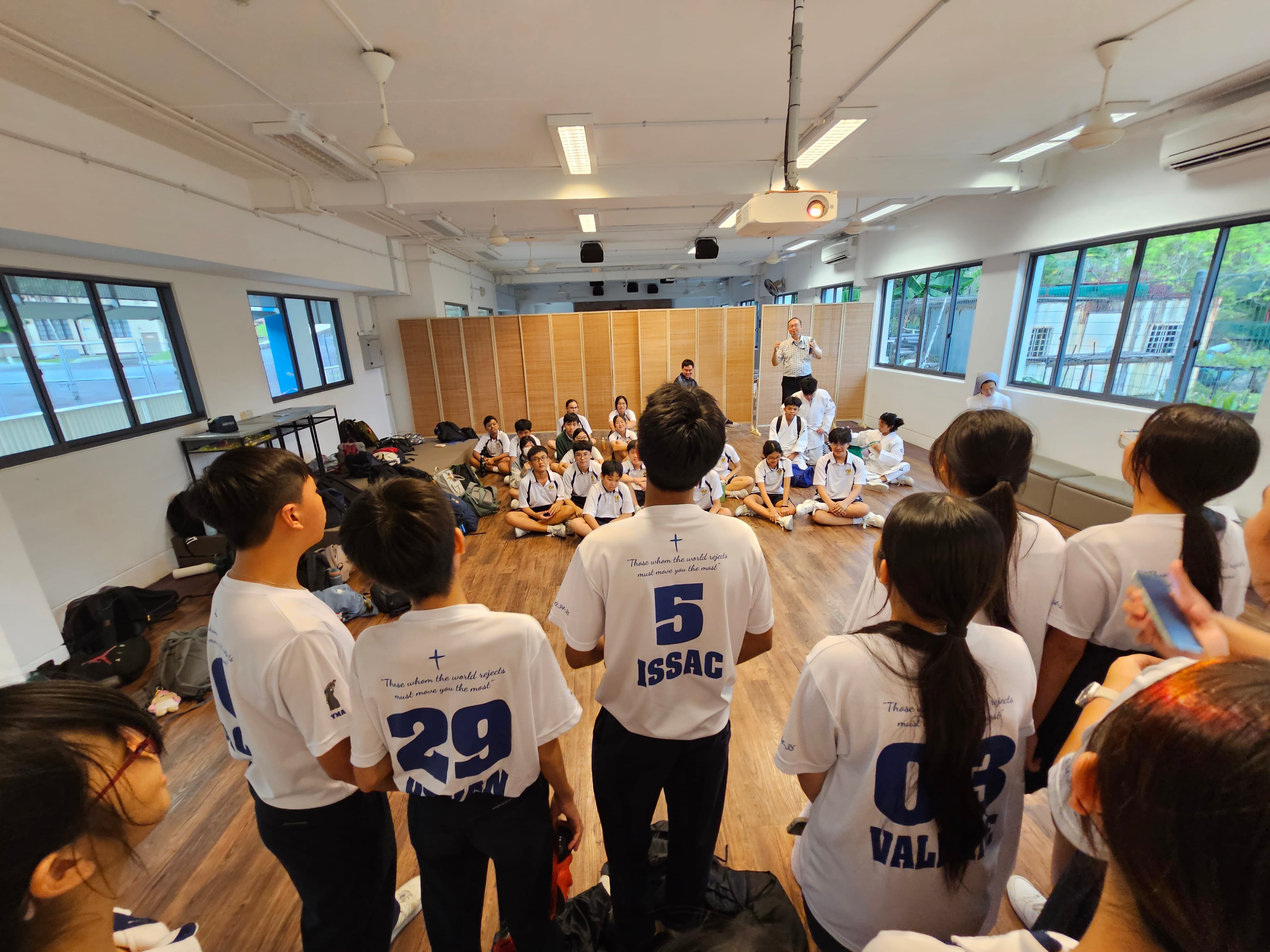 Students in numbered white jerseys stand and sit on a wooden floor in a bright room while an adult speaks at the front.
