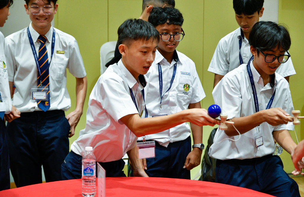 Students in white uniforms with lanyards play with kendama toys at a red table while others watch.