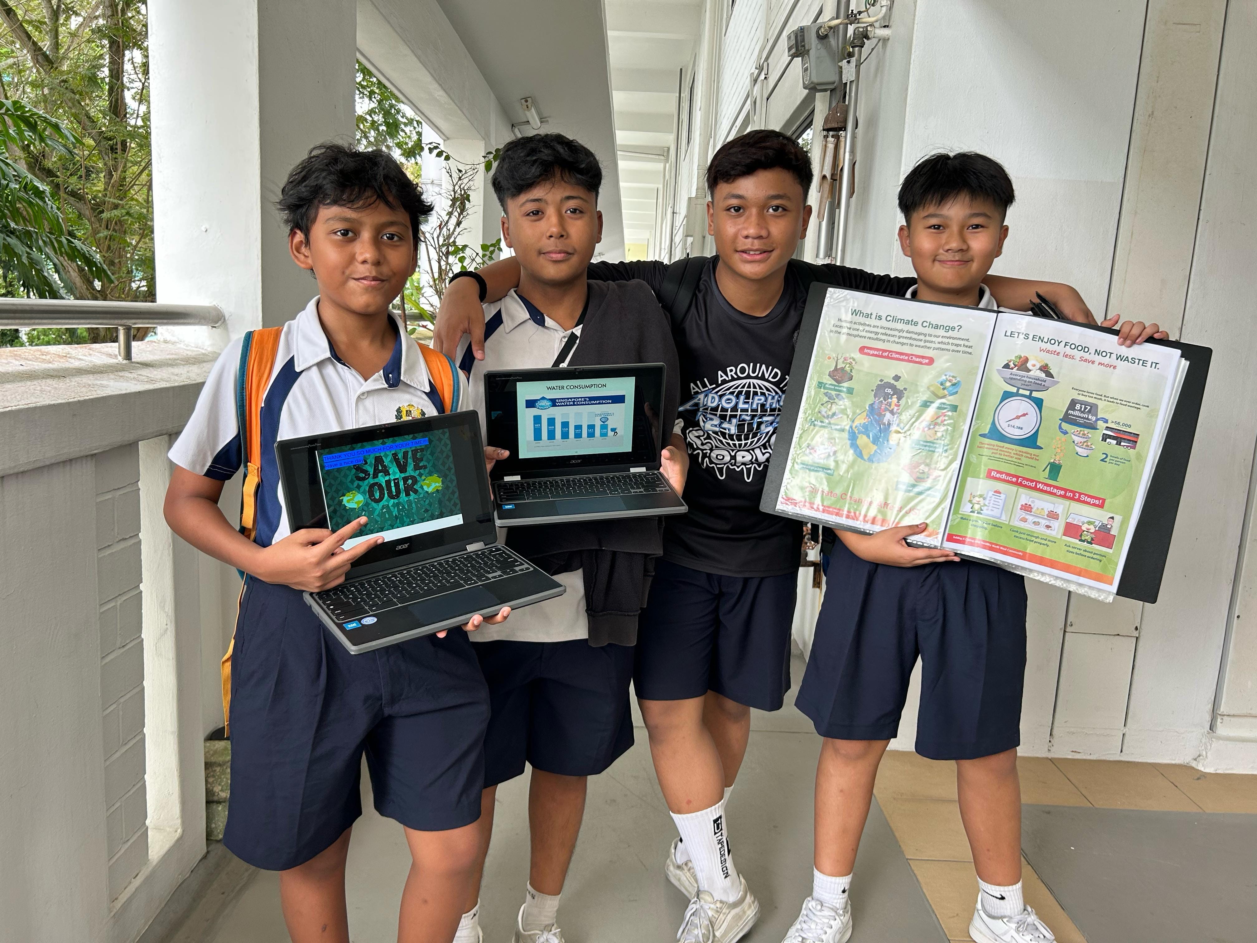 Four school boys stand in a corridor holding laptops showing environmental slides and a poster about climate change.