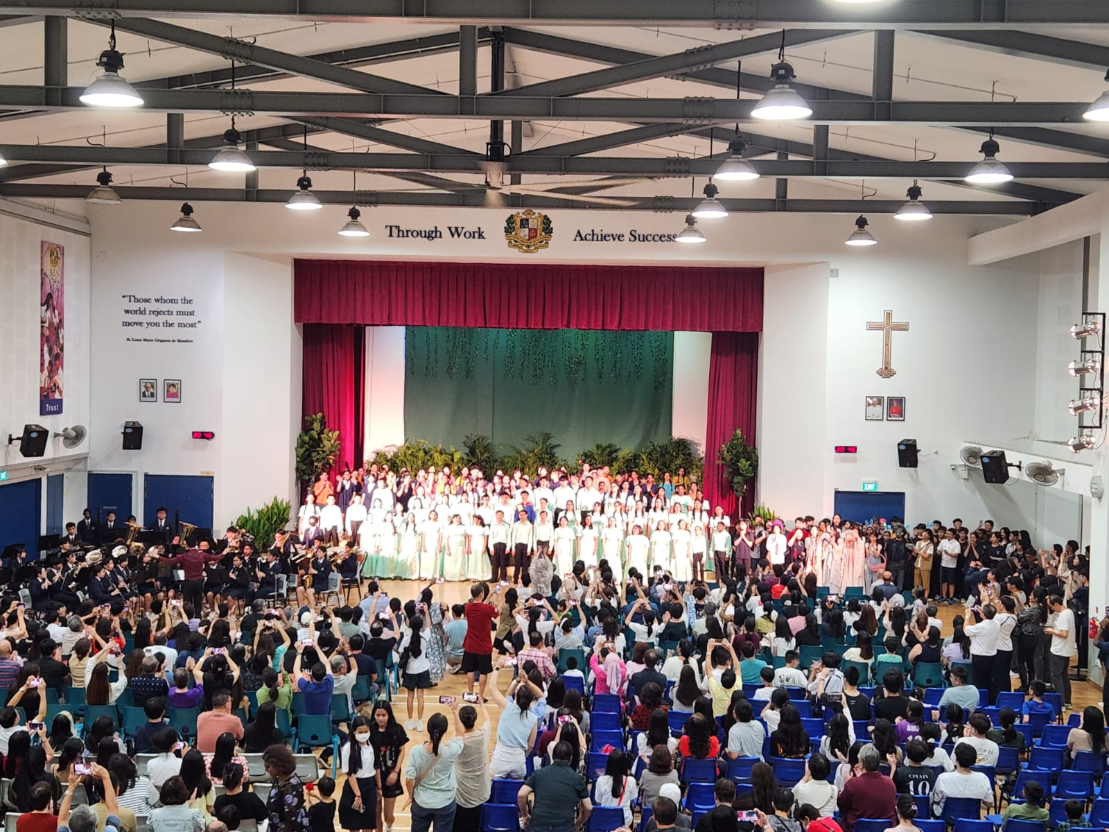 Crowded school hall with audience filming a large choir on stage under red curtains and bright lights.