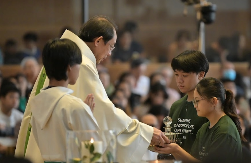 A priest in white vestments receives a glass vessel from two young people in green shirts during a church ceremony.