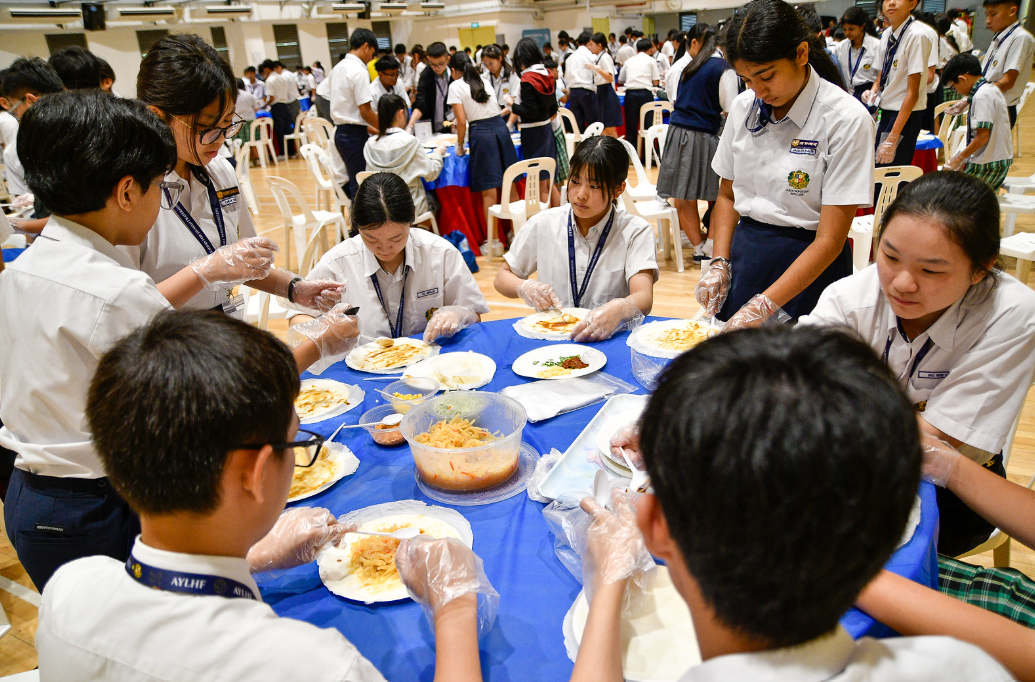 Students in white uniforms wearing plastic gloves prepare food on plates around a blue-covered table in a large hall.