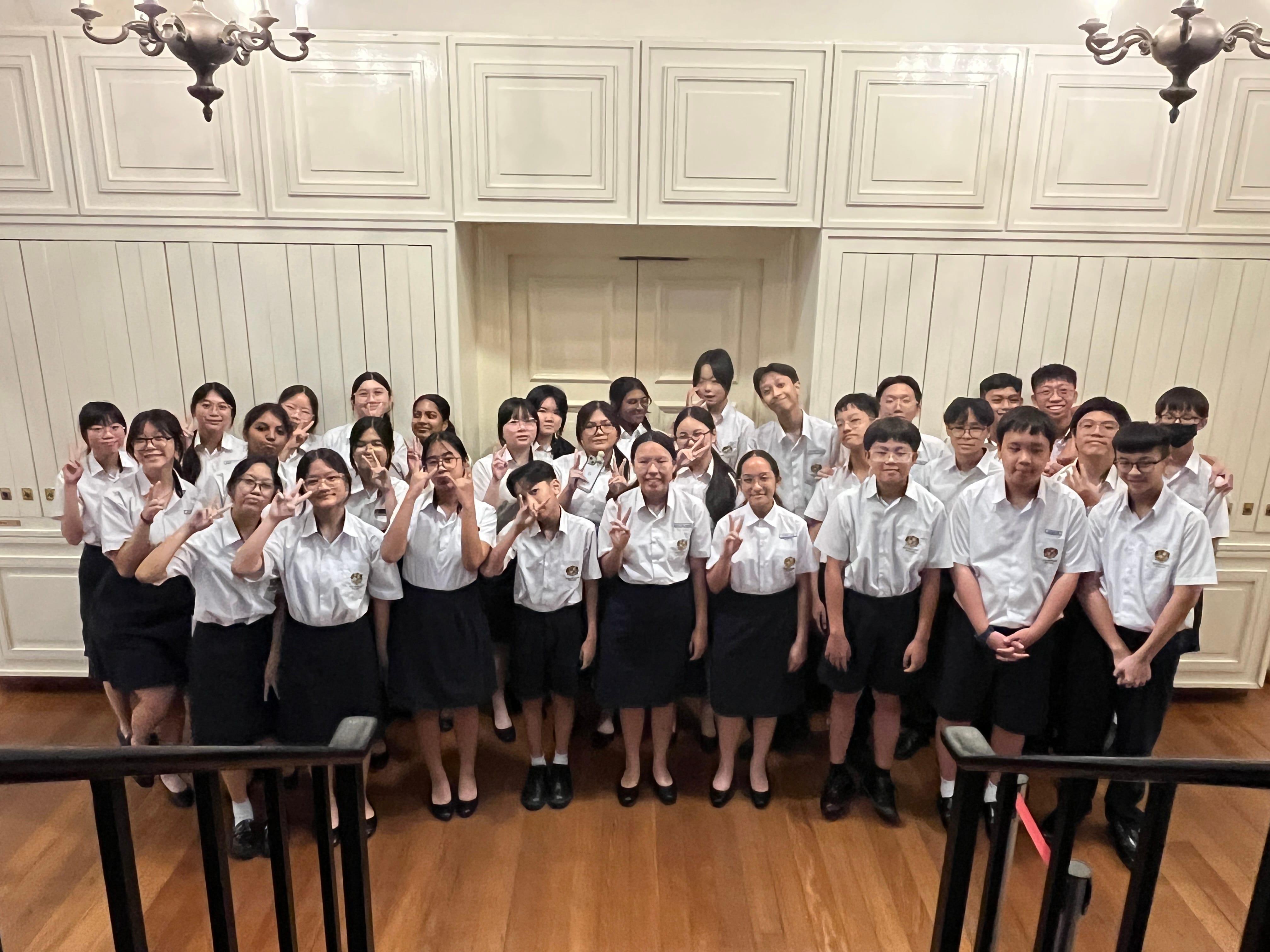 A large group of students in white shirts and dark skirts or pants pose with peace signs in an elegant room.