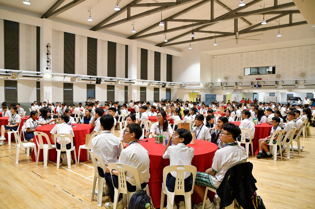 Students in white uniforms sit at round red-clothed tables in a large indoor hall during a school event.