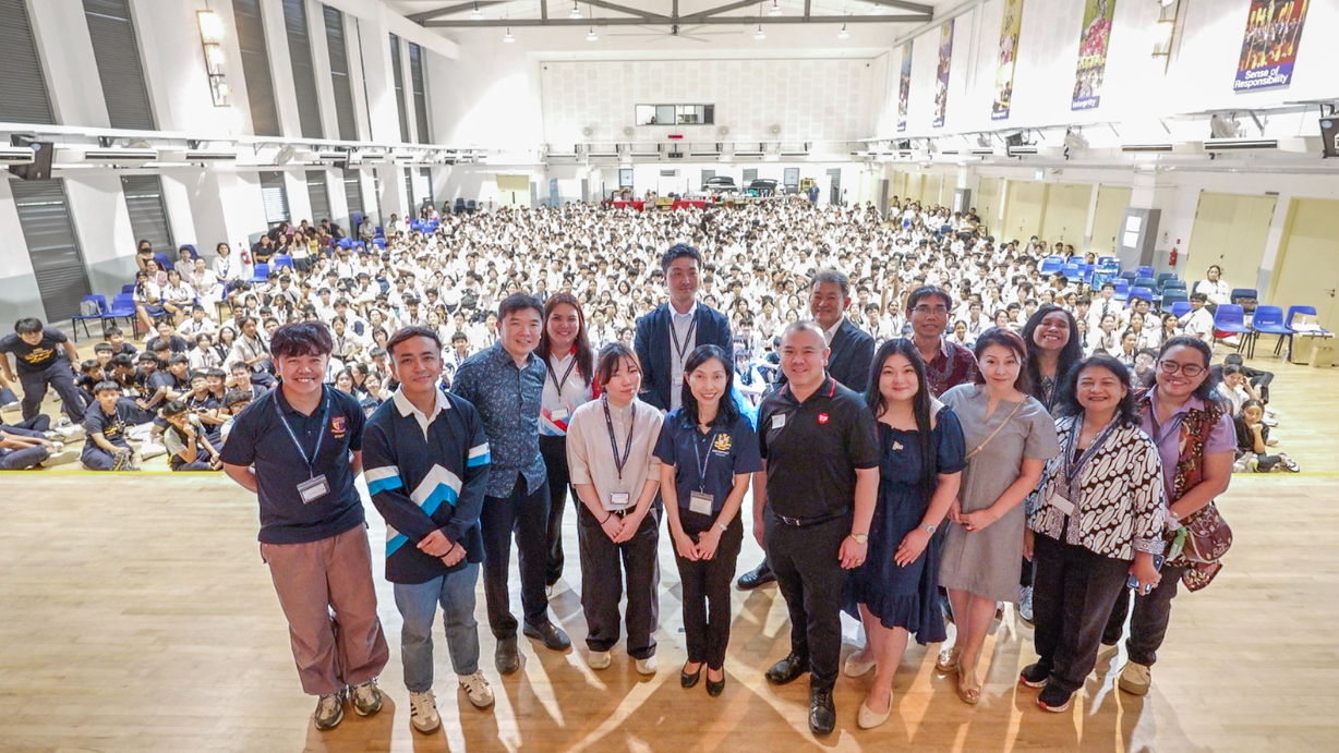 A group of adults poses in front of a large school hall filled with hundreds of students in white uniforms.