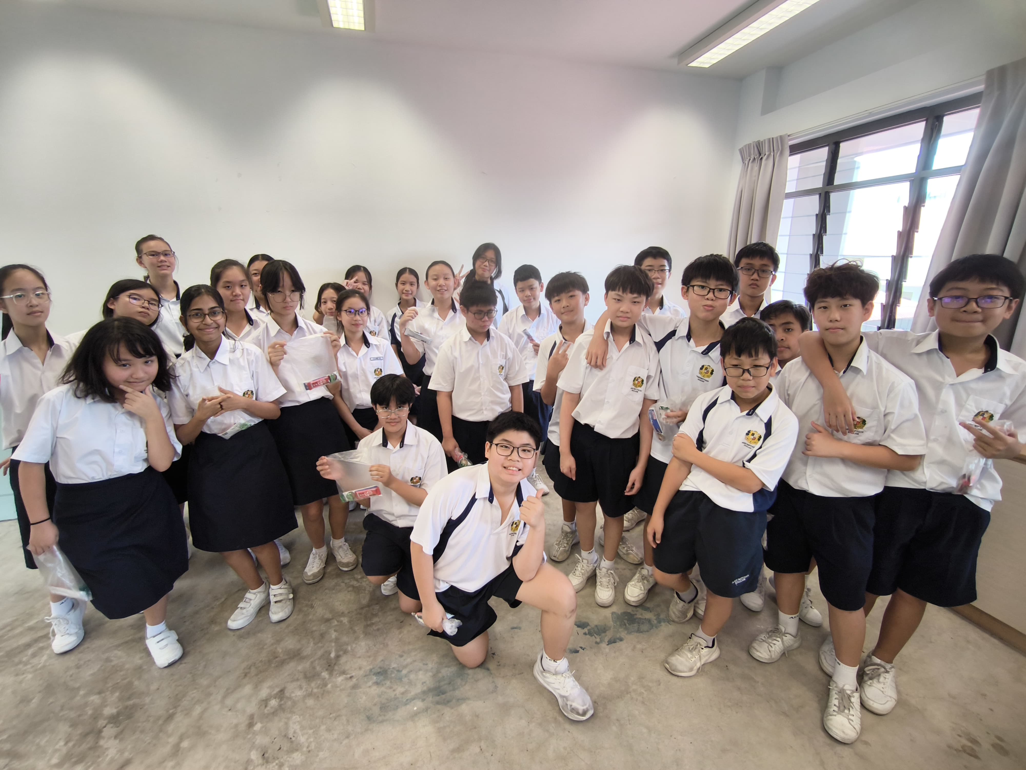 A large group of students in white school uniforms pose together smiling in a bright classroom.