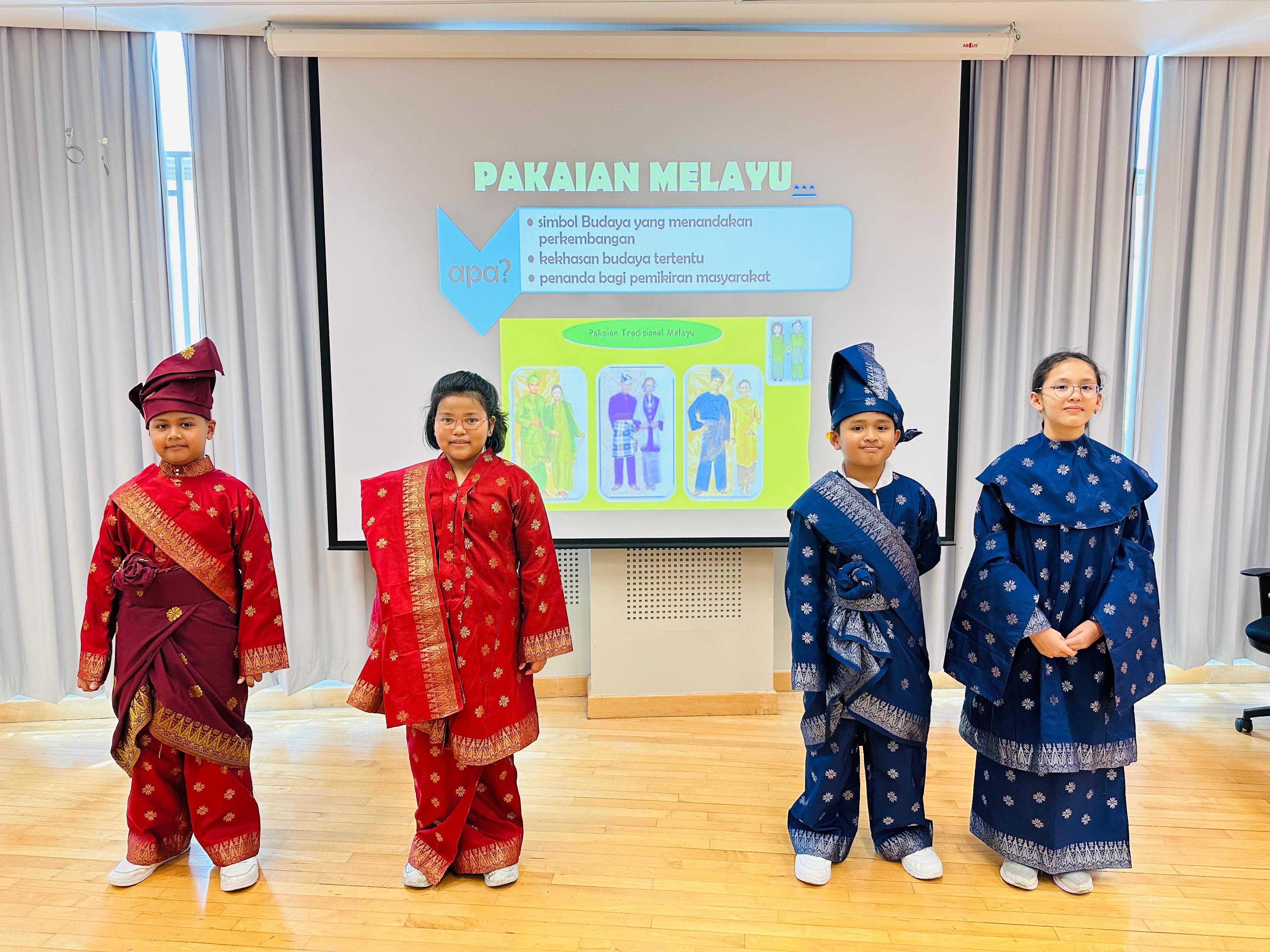 Four students standing side-by-side in traditional Malay clothing on stage in front of a presentation slide titled "PAKAIAN MELAYU...", featuring two boys wearing tengkolok headgear and sampan wraps alongside two girls wearing embroidered attire in red and dark blue.