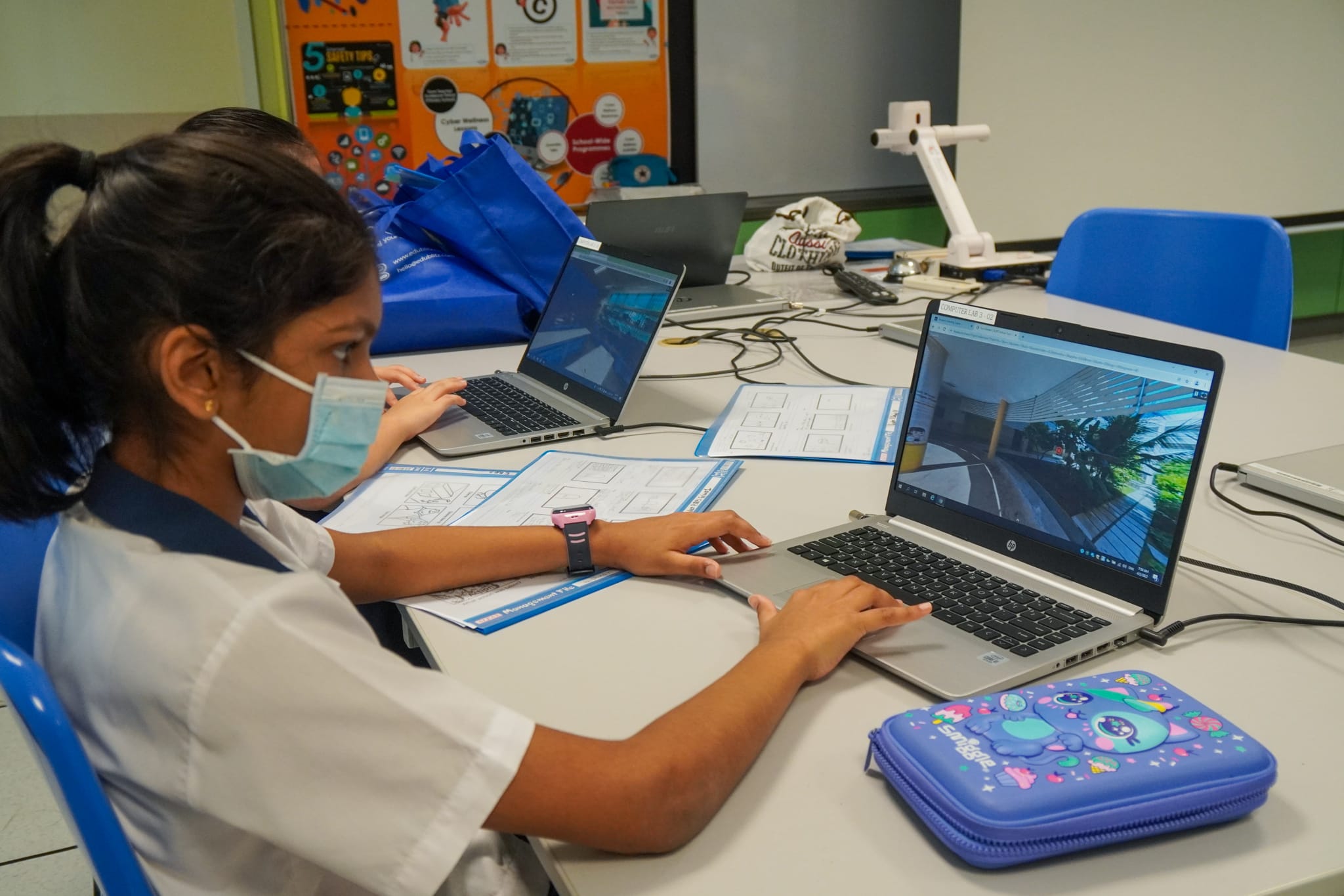 A masked schoolgirl in uniform works on a laptop at a classroom desk with papers and a pencil case nearby.