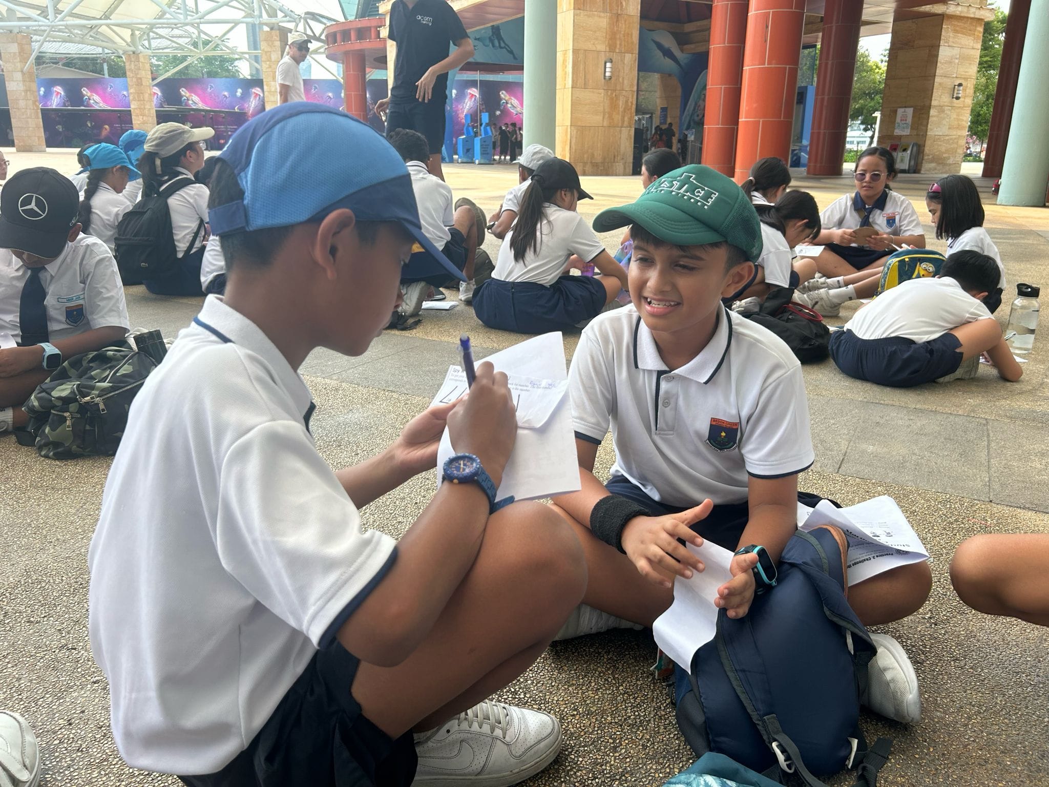 Two boys in school uniforms and caps sit on the ground outdoors, one writing on paper while the other smiles.