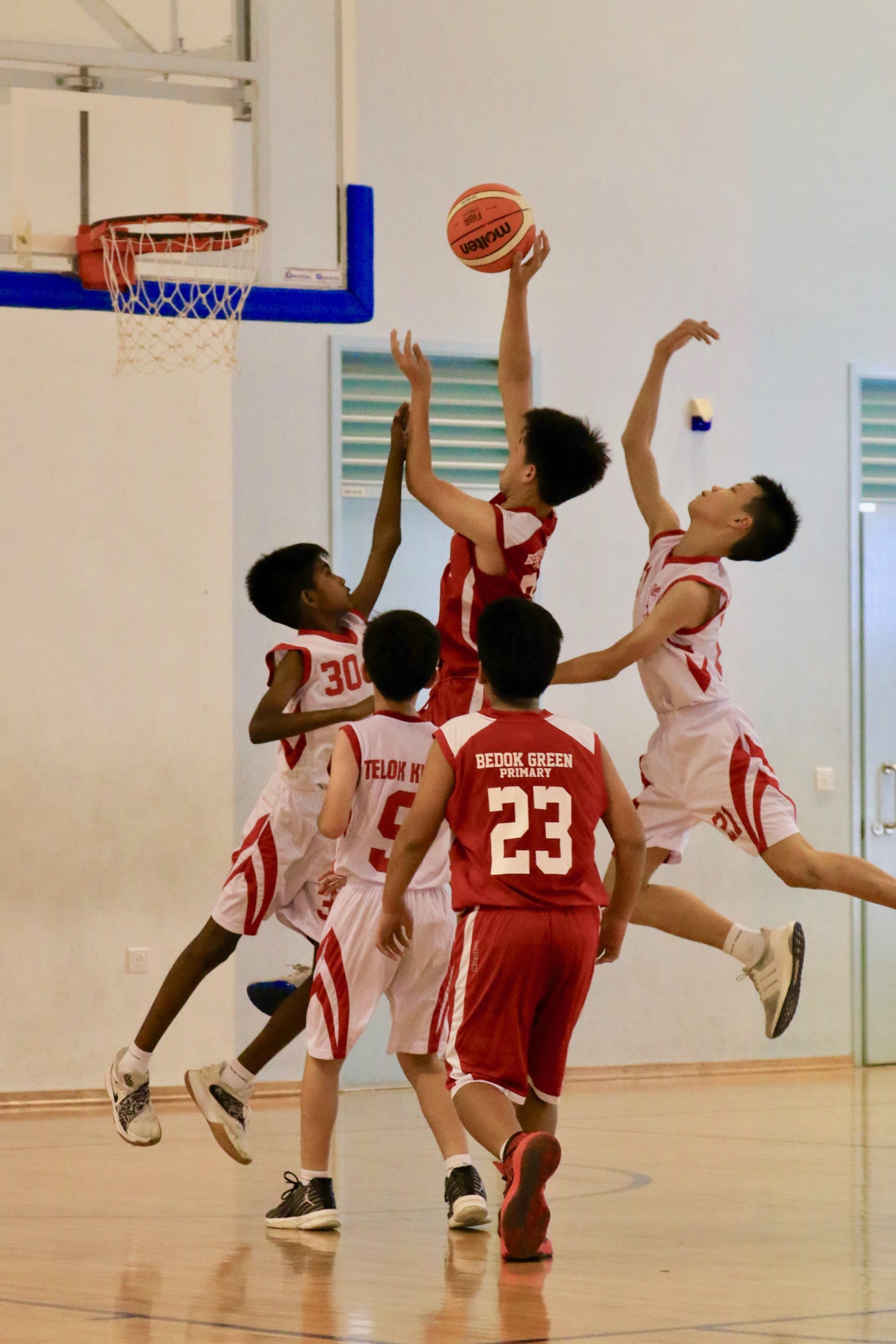 Young boys in red and white jerseys compete near a basketball hoop, with one player leaping to shoot the ball.