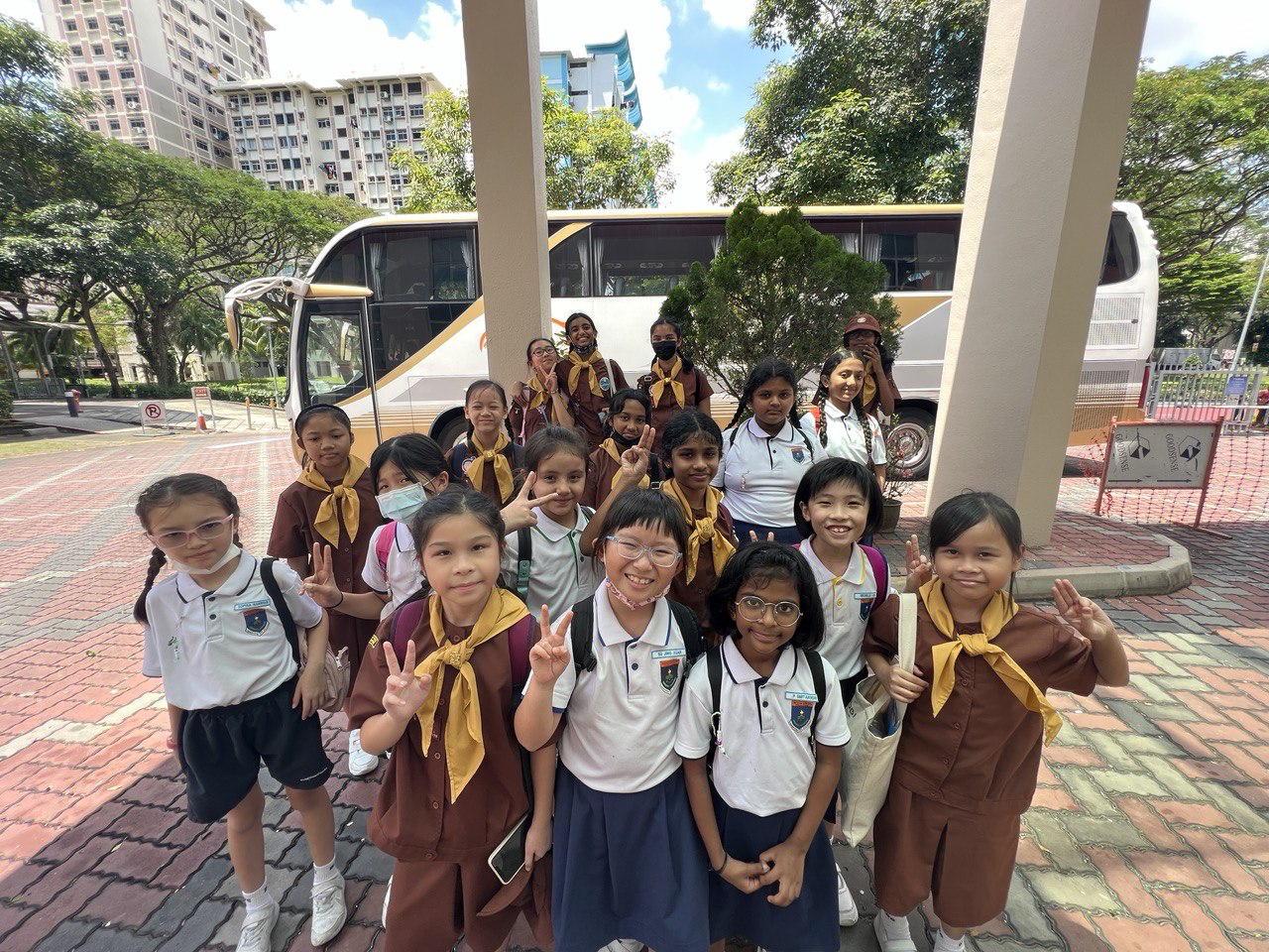 A group of young schoolgirls in uniforms pose with peace signs in front of a white coach bus outdoors.