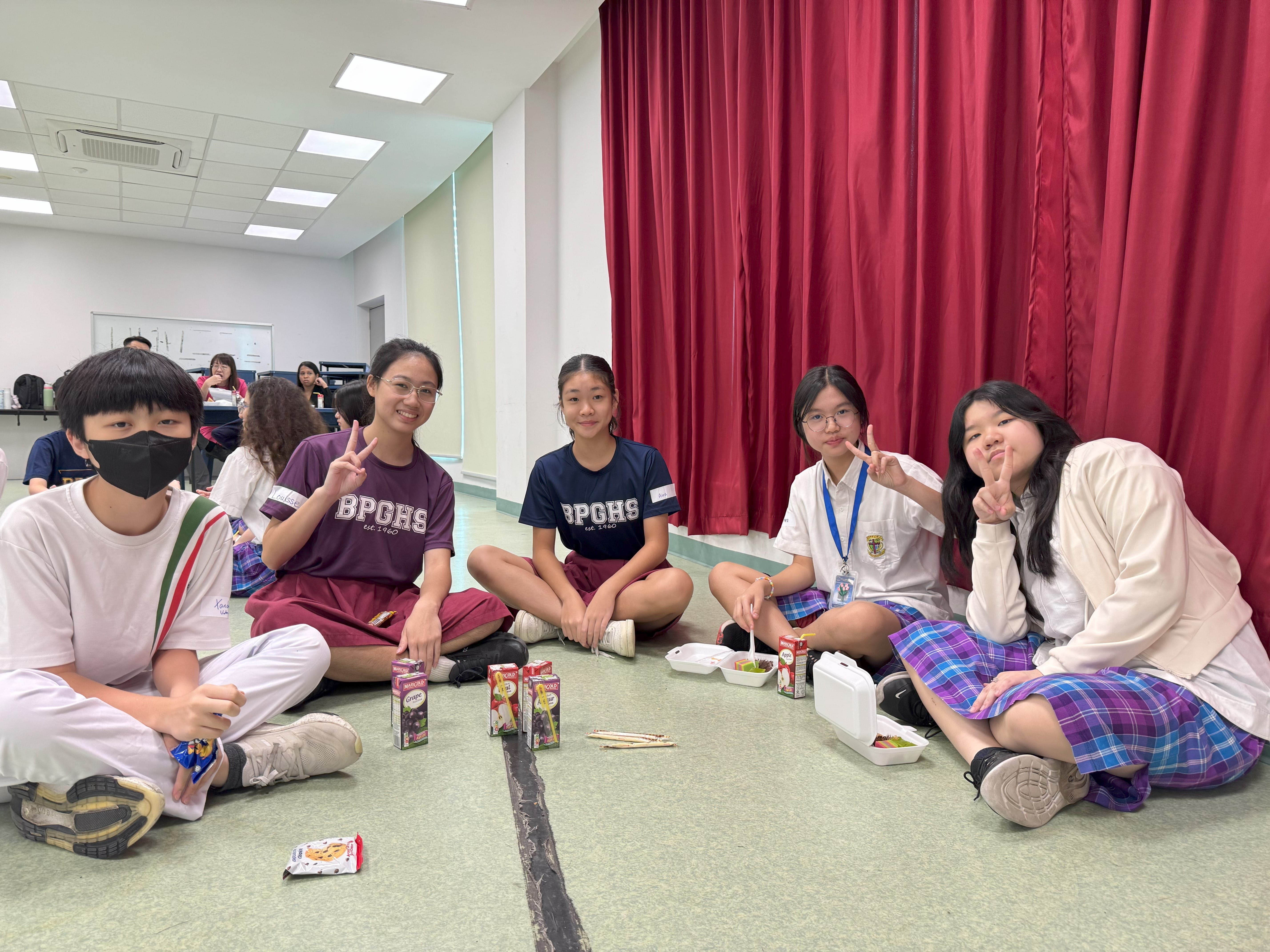 Five students in school uniforms sit on a floor near a red curtain, smiling and making peace signs.