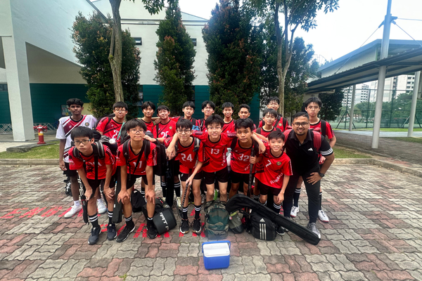 A youth field hockey team in red jerseys poses outdoors on a brick path with trees and buildings behind them.