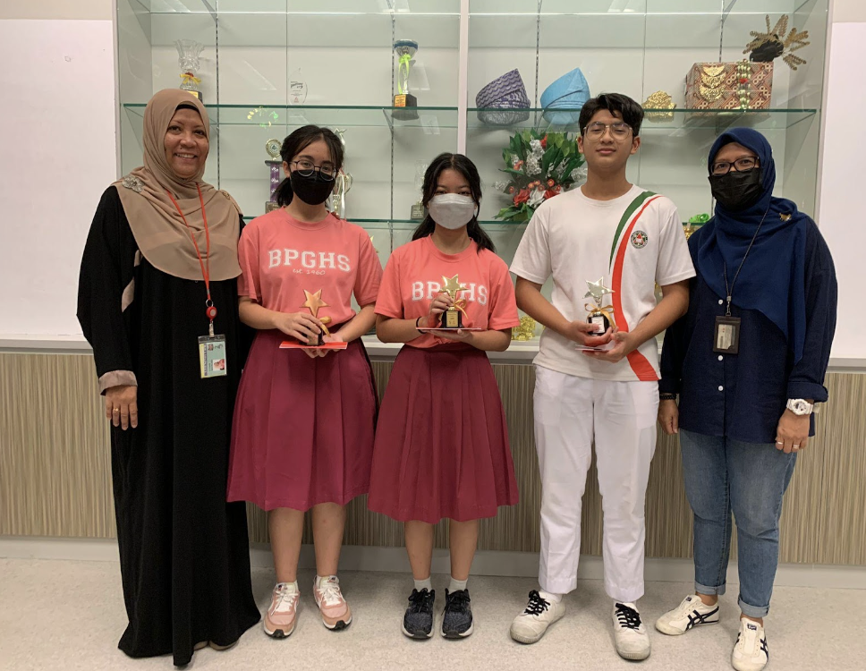Five people pose together indoors, with three students holding star-shaped trophies, flanked by two adults wearing hijabs.