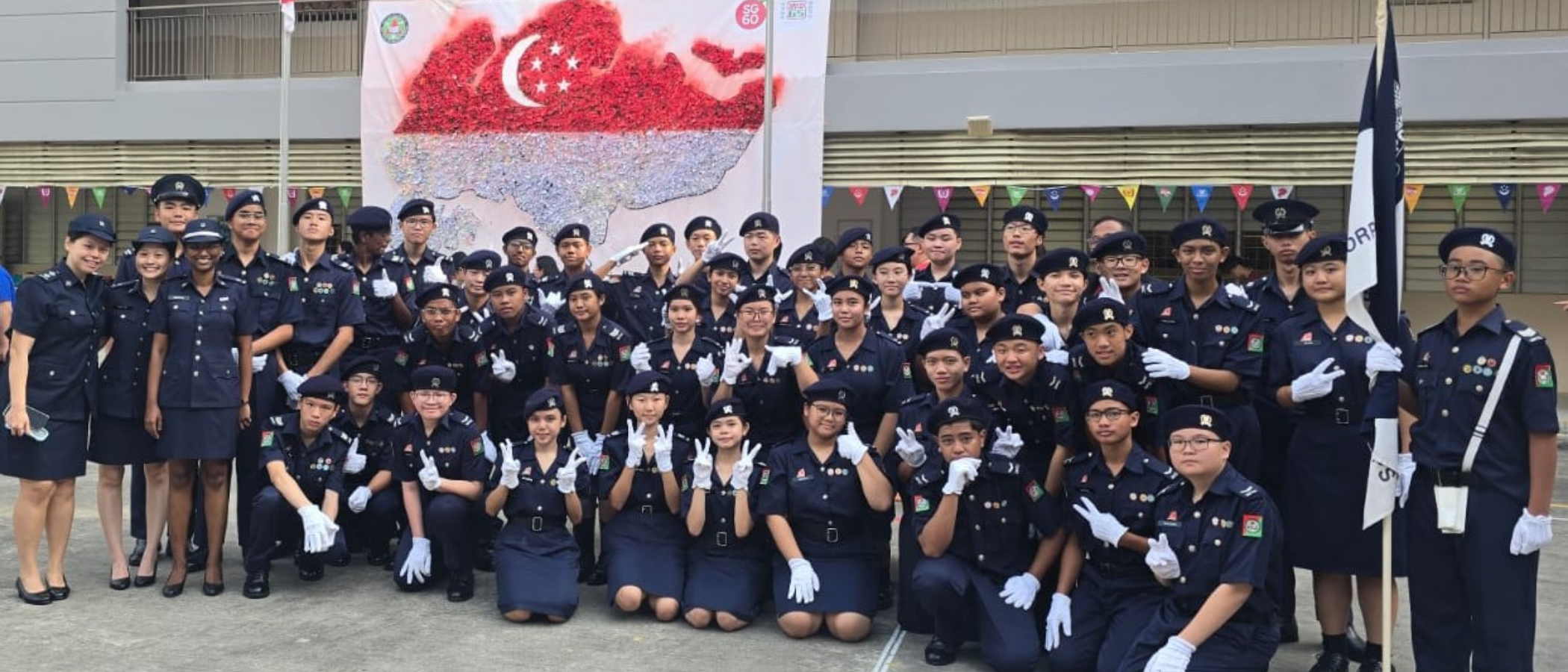 Large group of youth in dark navy uniforms and white gloves posing outdoors before a Singapore flag banner.