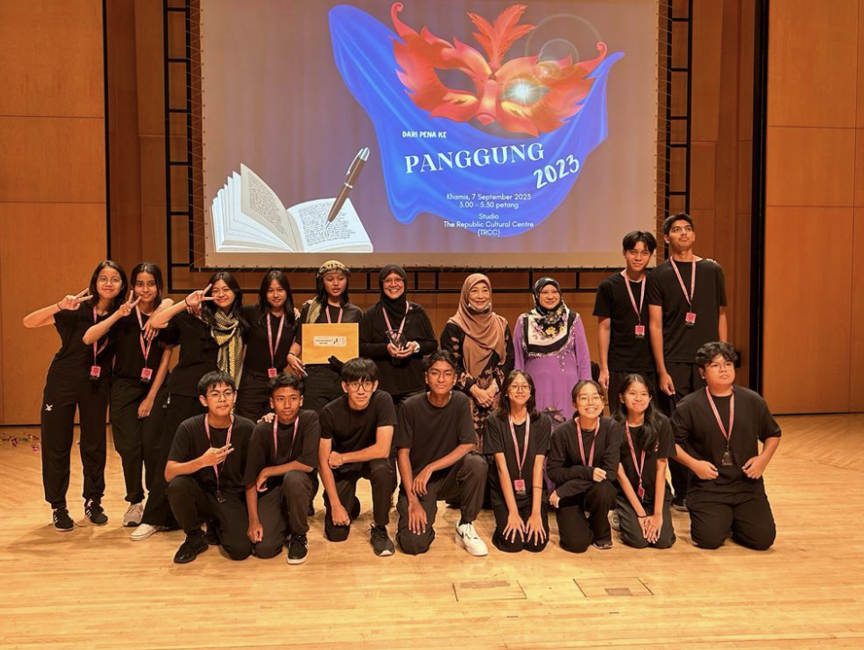 A group of young performers in black outfits pose on stage before a Panggung 2023 event projection.
