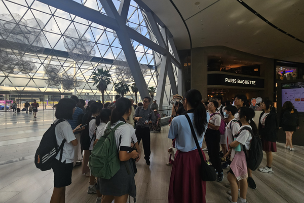 A group of students listens to a guide inside a modern mall with a large geometric glass facade and a Paris Baguette store nearby.