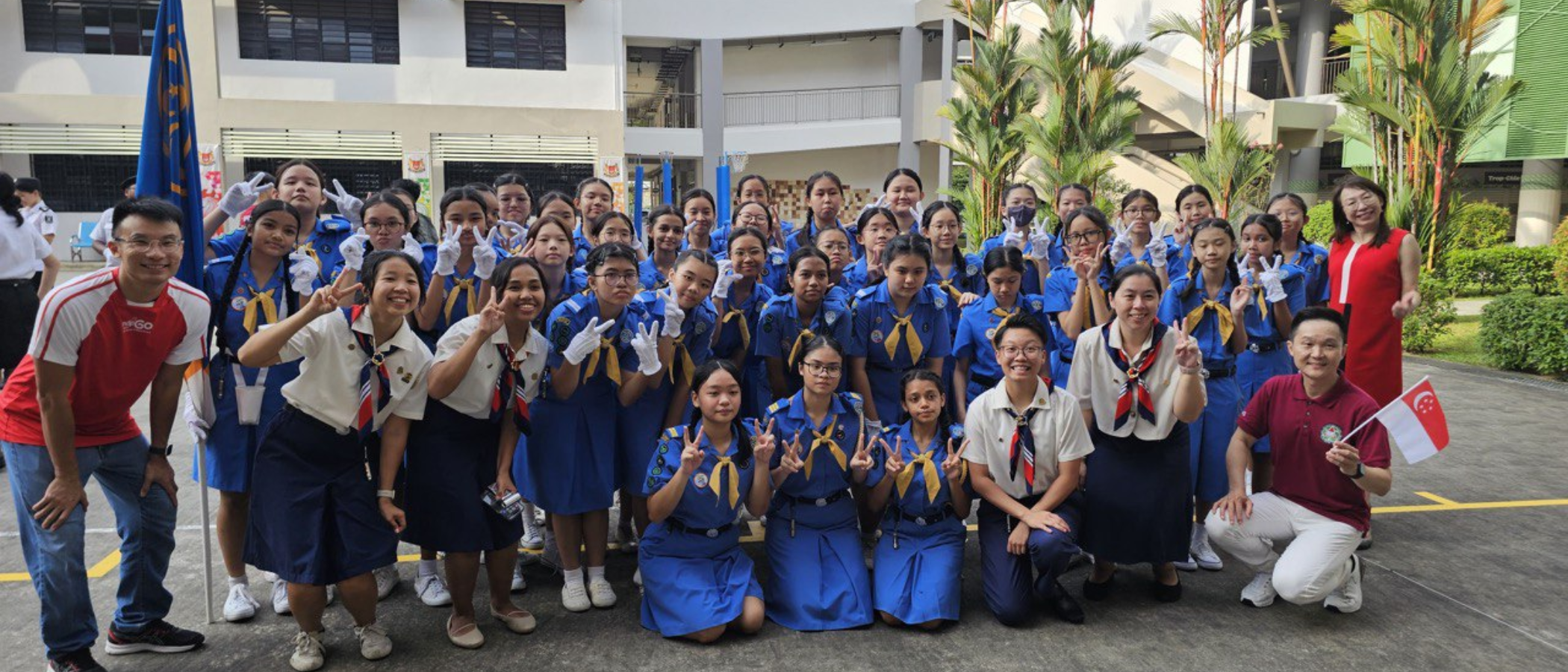 Large group of students in blue uniforms posing with peace signs outdoors at a school, one person holds a Singapore flag.