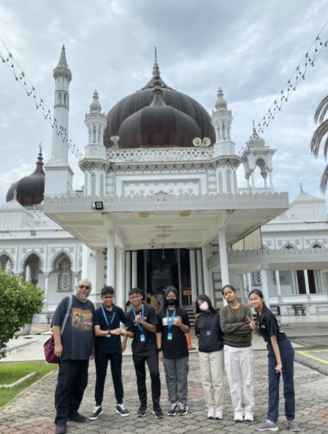 Six people stand in front of a white mosque with a large dark brown dome and minarets under a cloudy sky.
