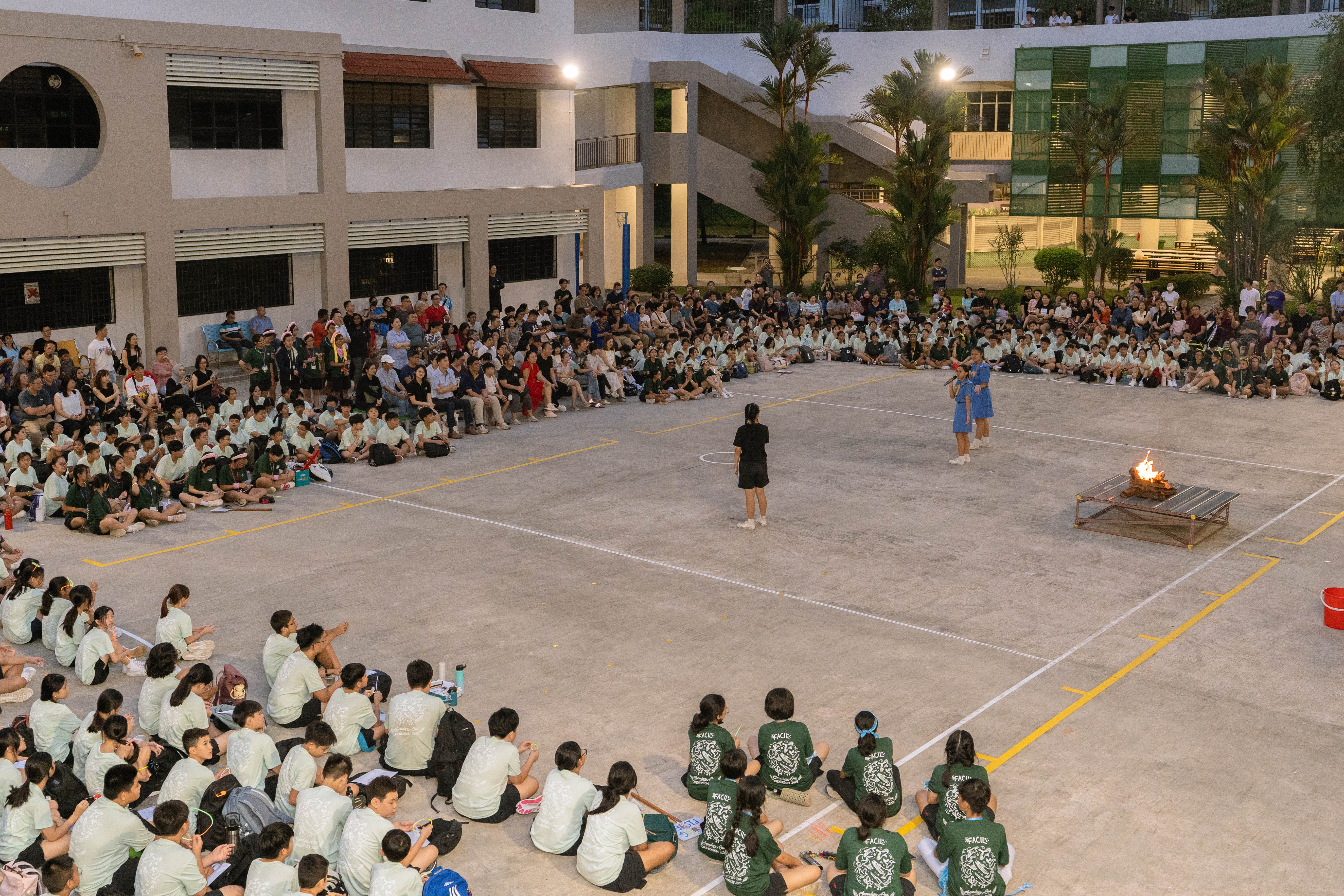 Students in uniforms sit around a school courtyard at dusk watching performers near a small bonfire.