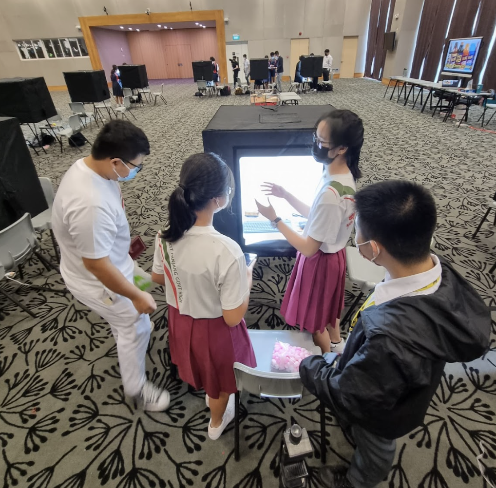 Four masked students in school uniforms gather around a glowing light box display in a large carpeted hall.