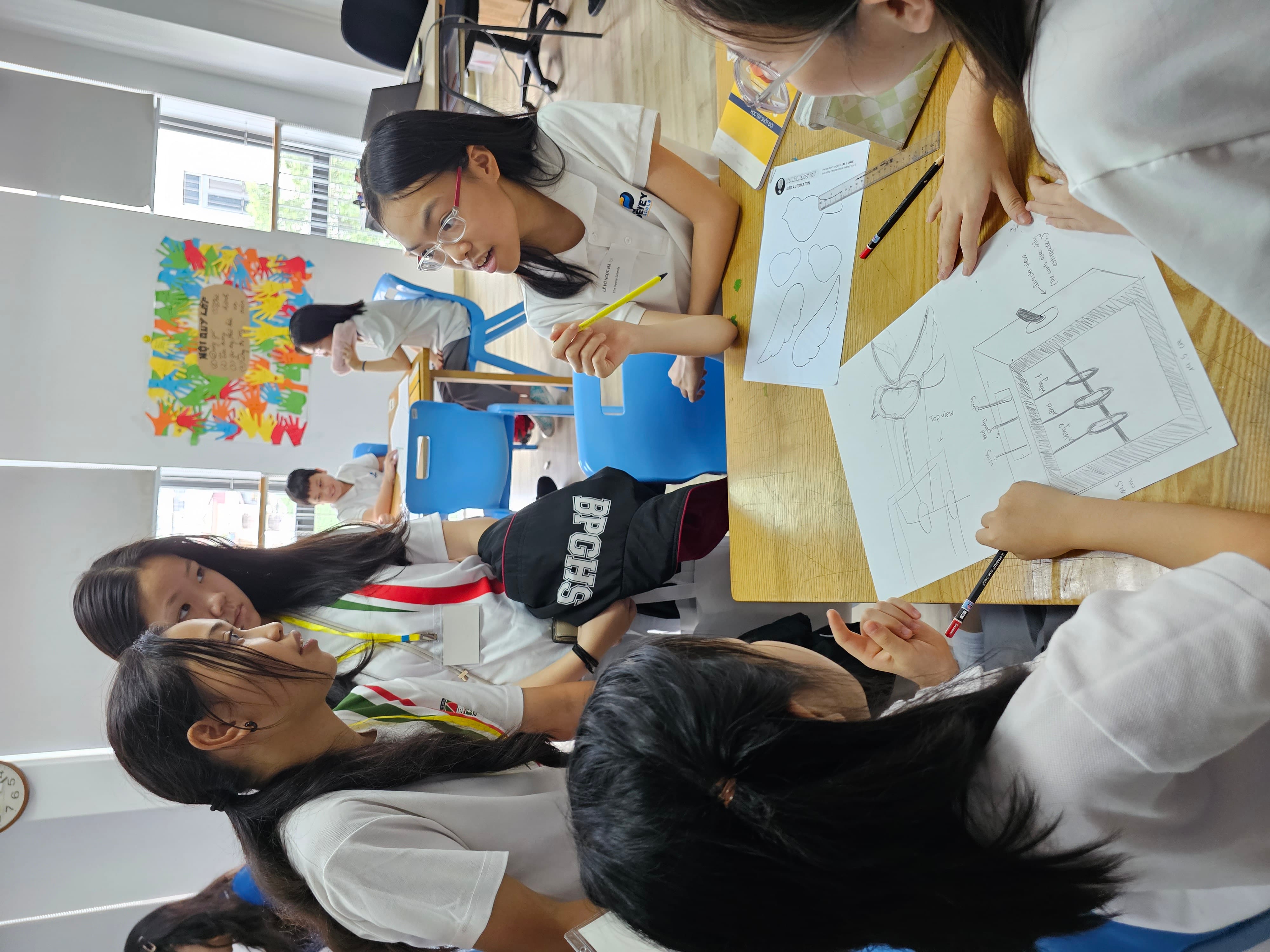 Several students in white uniforms gather around a desk reviewing pencil sketches and drawings in a bright classroom.