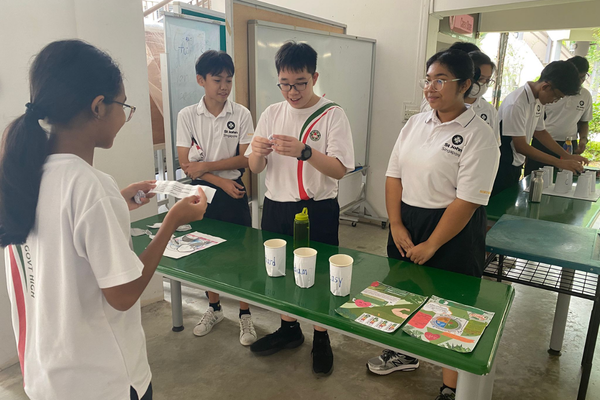 Four students in white uniforms stand around a green table with paper cups and printed materials during a school activity.