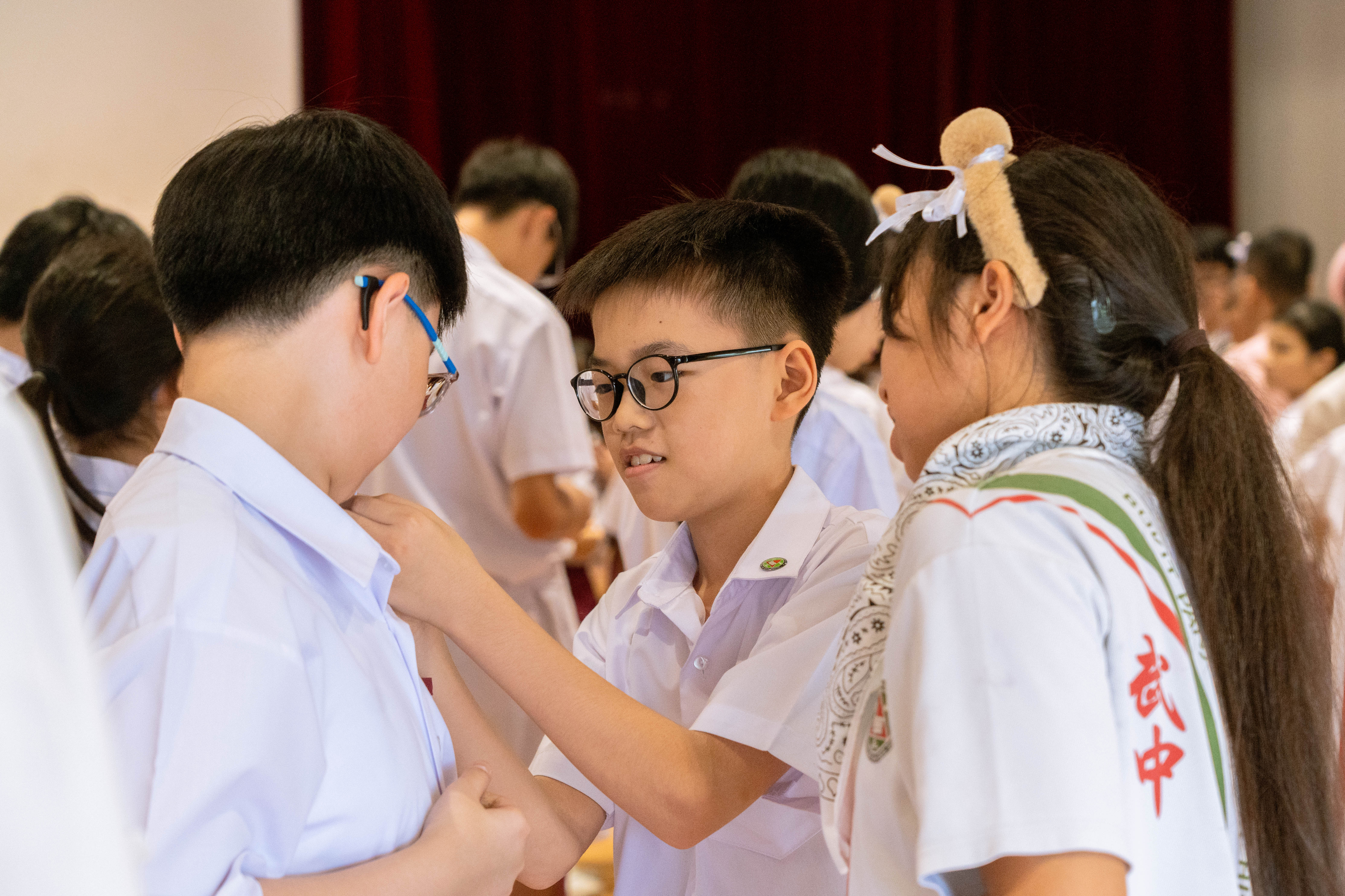 Three young students in white uniforms interact closely, with a boy wearing glasses reaching toward another student.