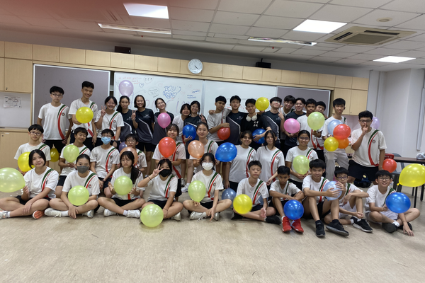 Group of students in white uniforms posing in a classroom, holding colorful balloons in yellow, red, and blue.