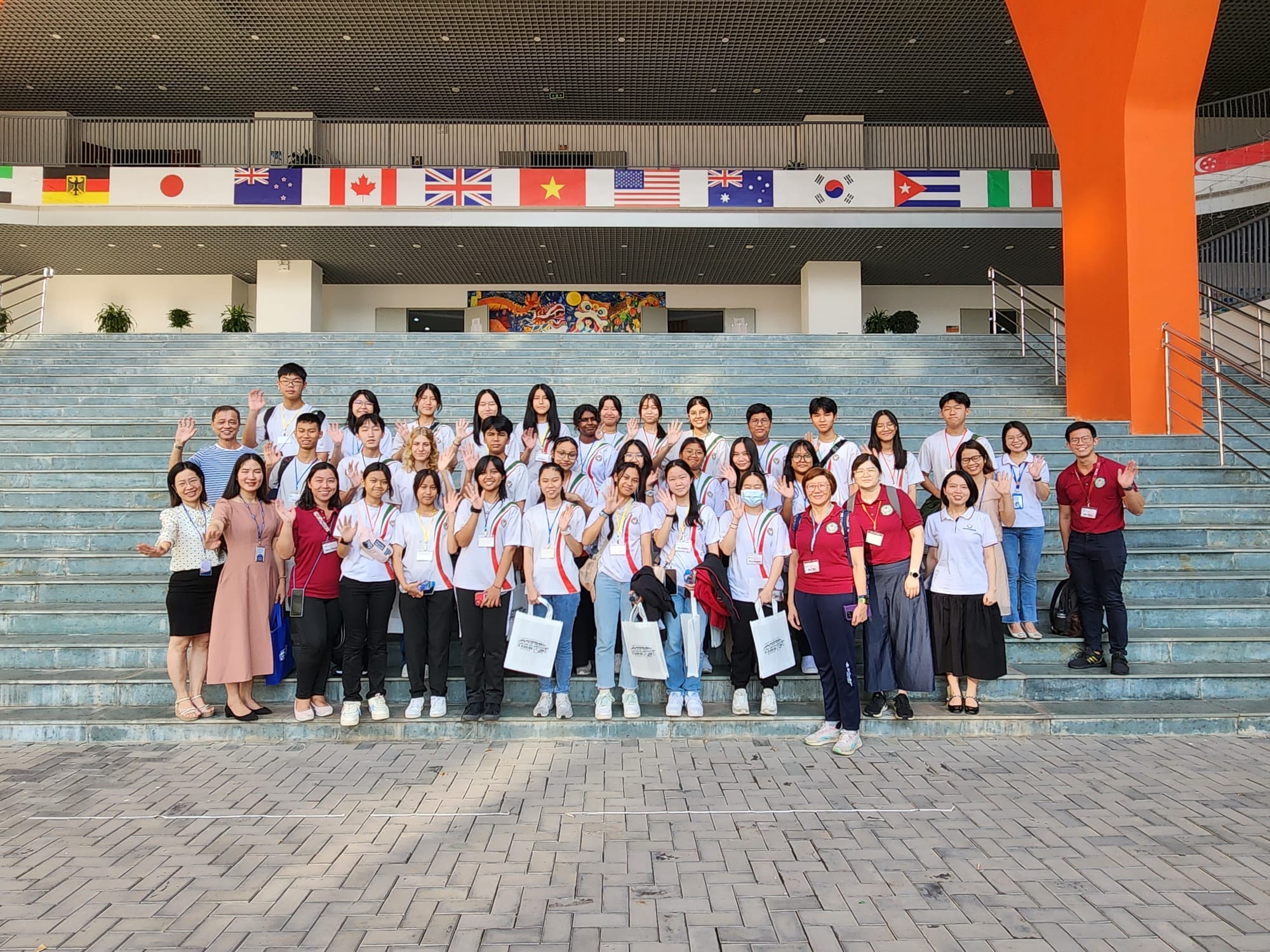 A large group of students and teachers pose on outdoor steps of a building decorated with international flags.