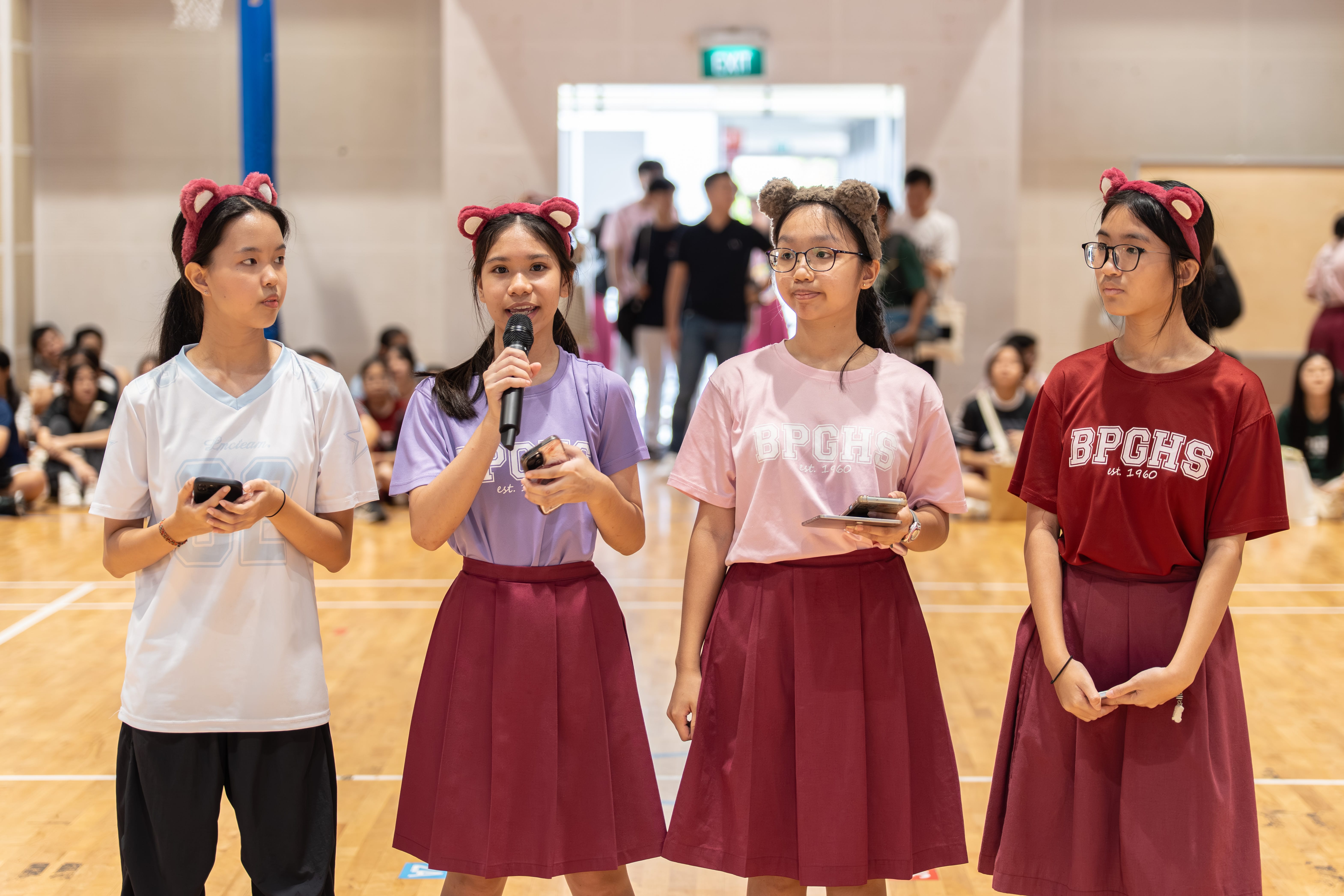Four girls in BPGHS uniforms with bear ear headbands stand in a gymnasium, one speaking into a microphone.