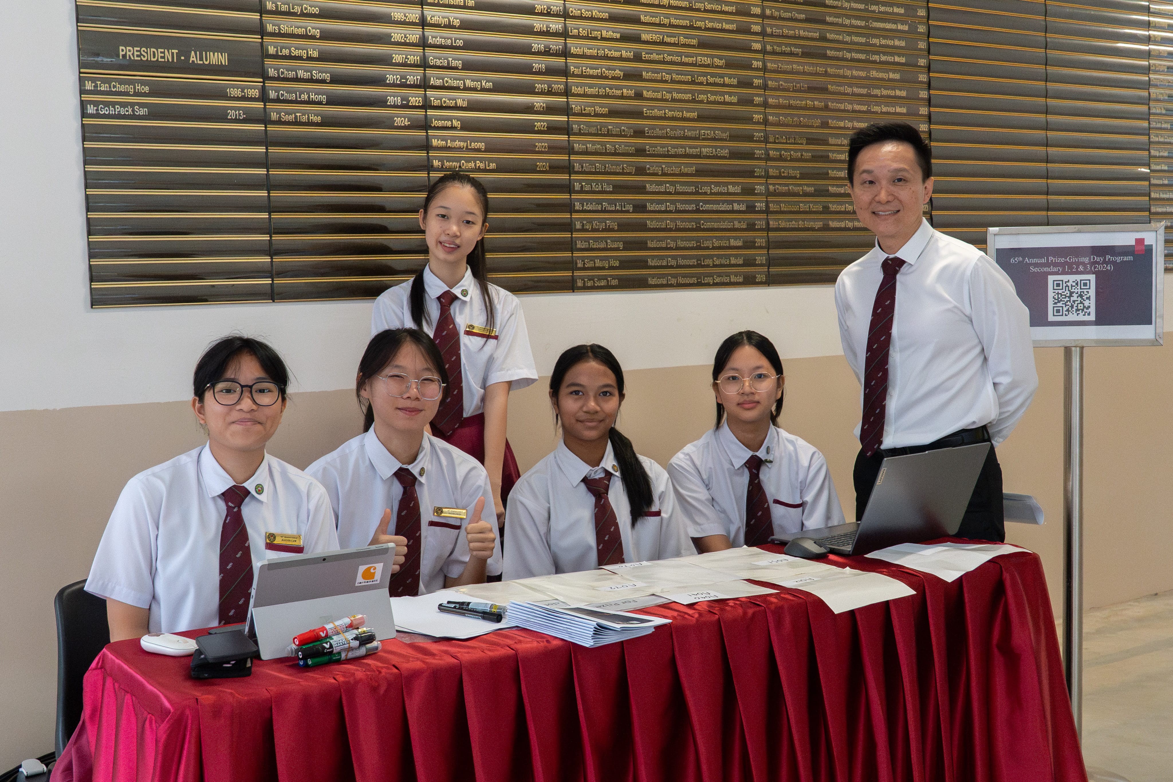 Five students in white uniforms and a man in a shirt and tie pose at a red-draped registration table indoors.