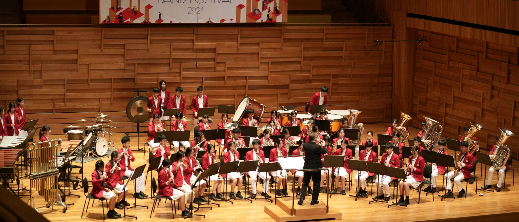 Youth band in red jackets performing on a concert hall stage at Band Festival 2024, led by a conductor.