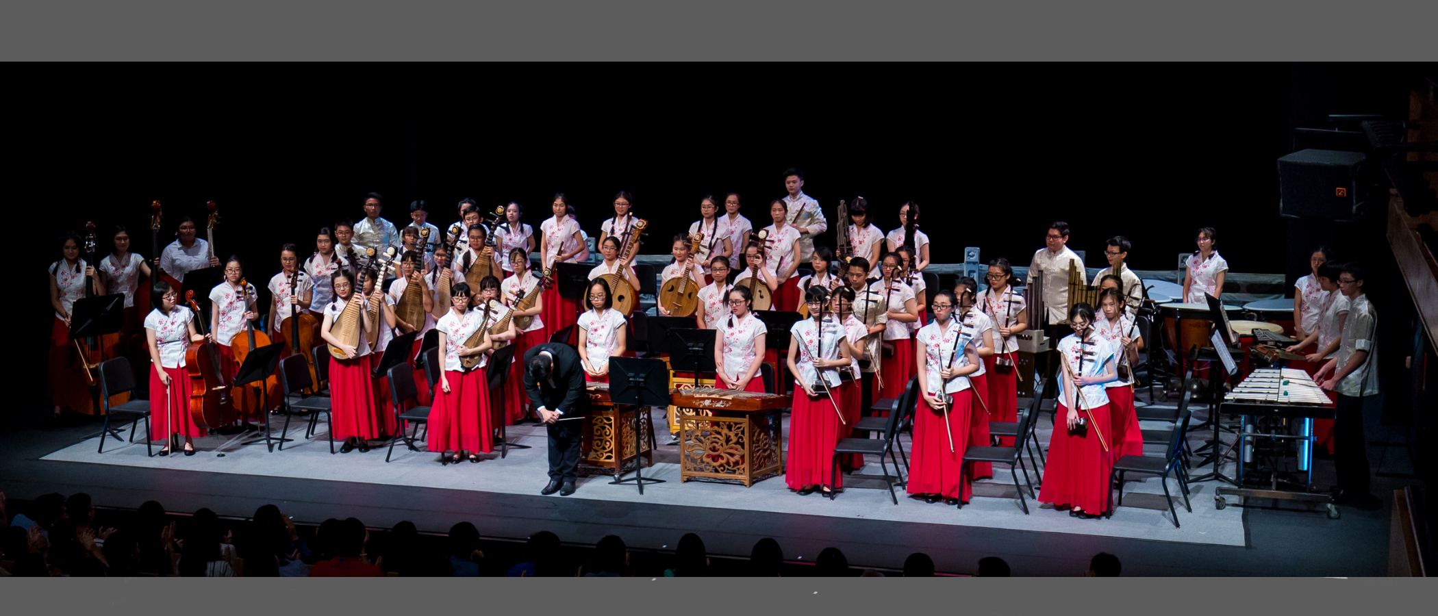 A large Chinese orchestra on stage, musicians in white tops and red skirts bowing after a performance.