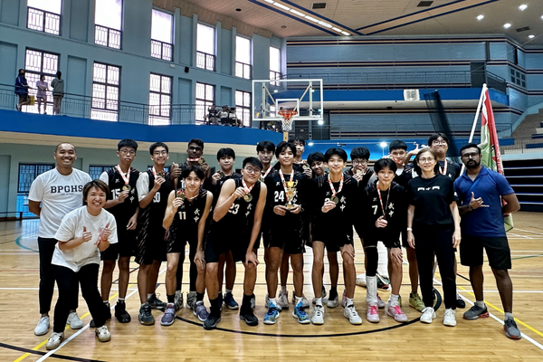A youth basketball team in black uniforms poses with medals and a trophy on an indoor court with coaches.