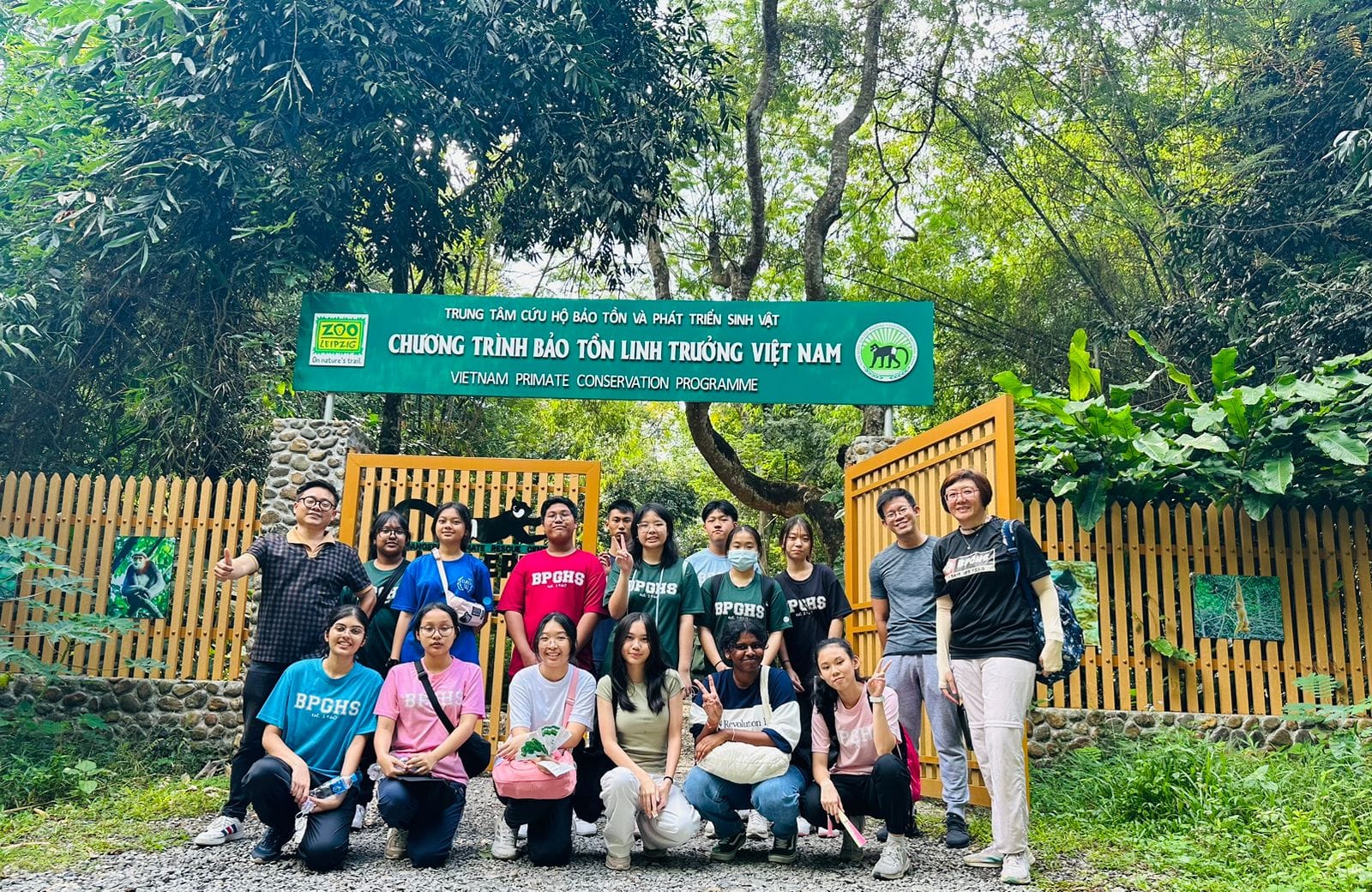 A group of students pose at the entrance gate of the Vietnam Primate Conservation Programme surrounded by trees.