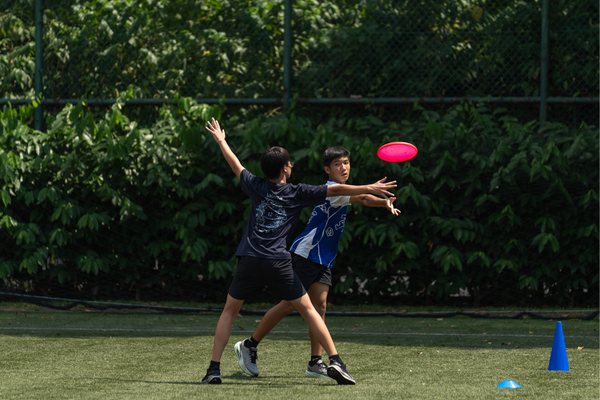 Two boys play ultimate frisbee on a grass field, one reaching to catch a red disc while the other defends.
