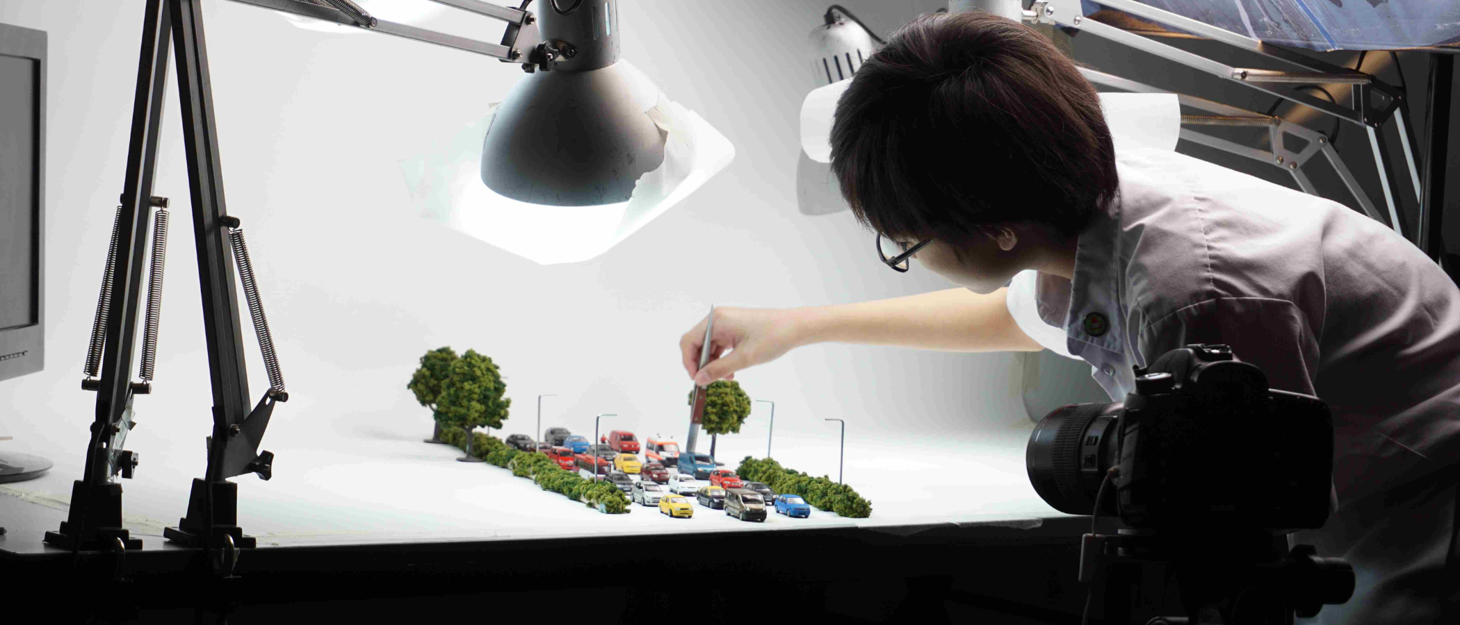 Person leaning over a lit table arranging tiny model cars and trees, with studio lamps and a camera nearby.