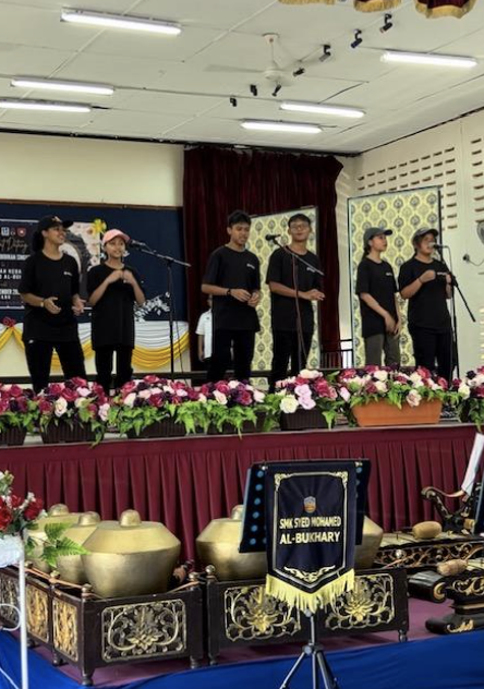 Six students in black uniforms stand on a stage with microphones, above traditional instruments and floral decorations.