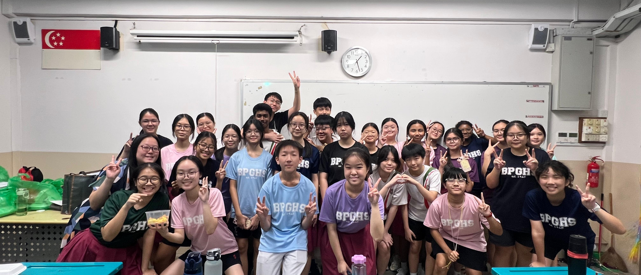 A large group of students in BPGHS shirts pose with peace signs in a classroom with a Singapore flag.