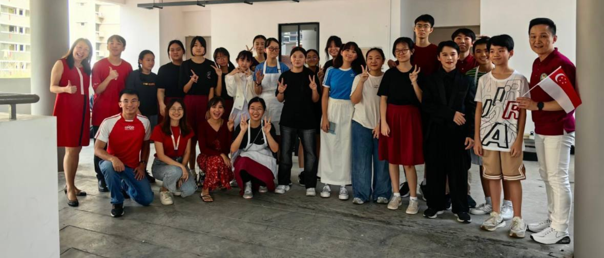 A large group of young people and adults posing together indoors, some making peace signs, one holding a Singapore flag.