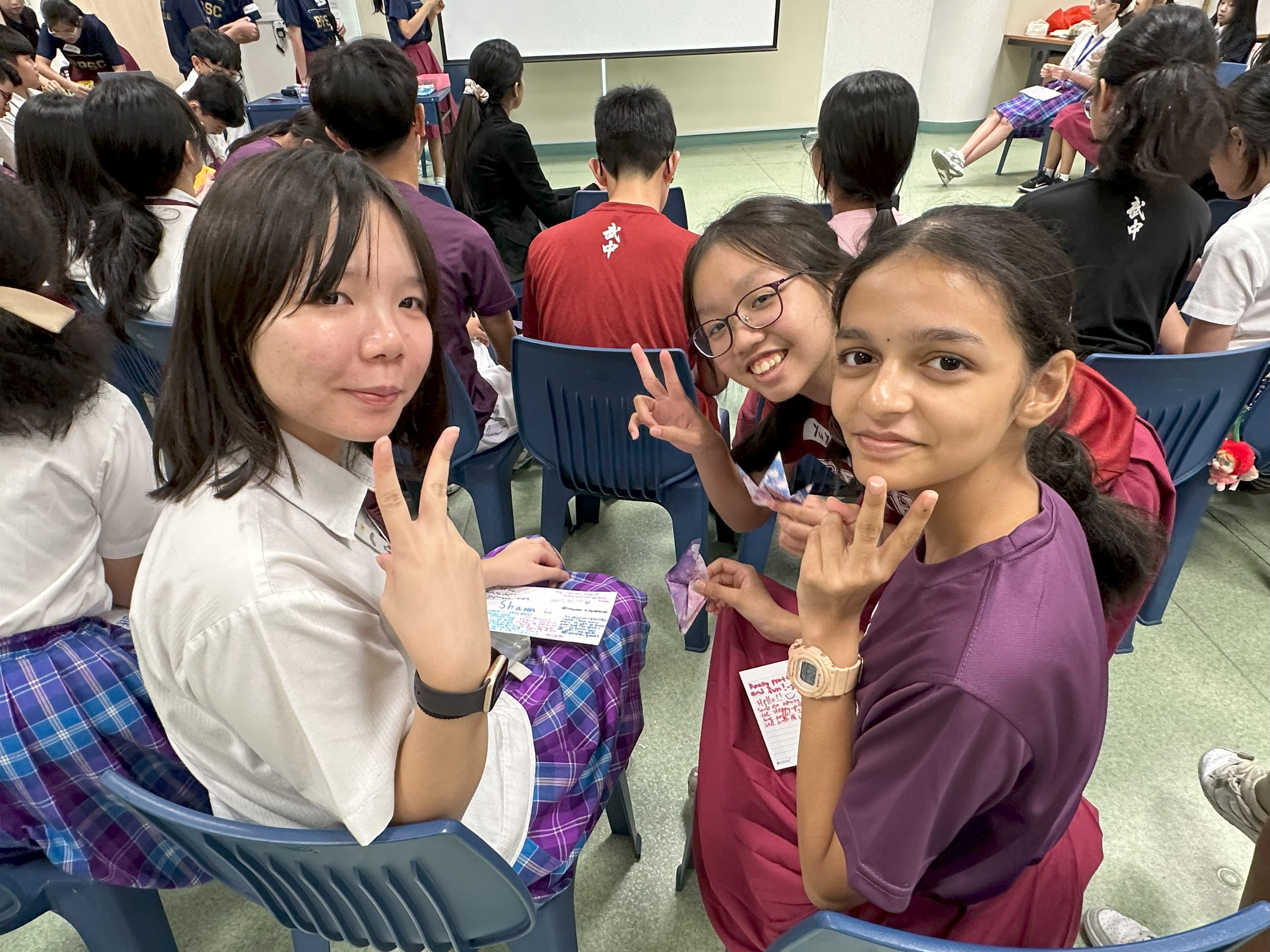 Three smiling schoolgirls pose with peace signs in a crowded classroom filled with other students.
